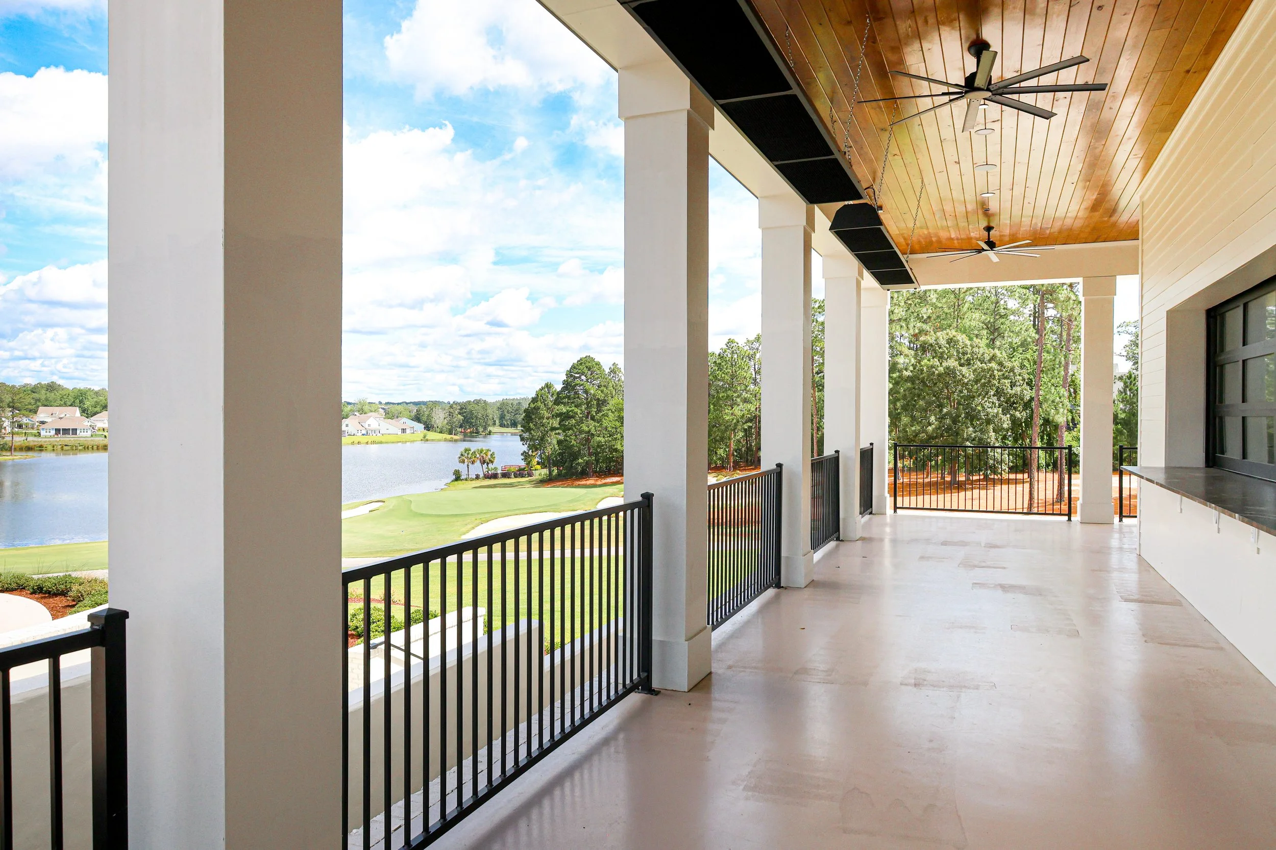 Outdoor balcony with black railing, ceiling fans, and a view of a pond, green grass, and trees under a blue sky with scattered clouds.