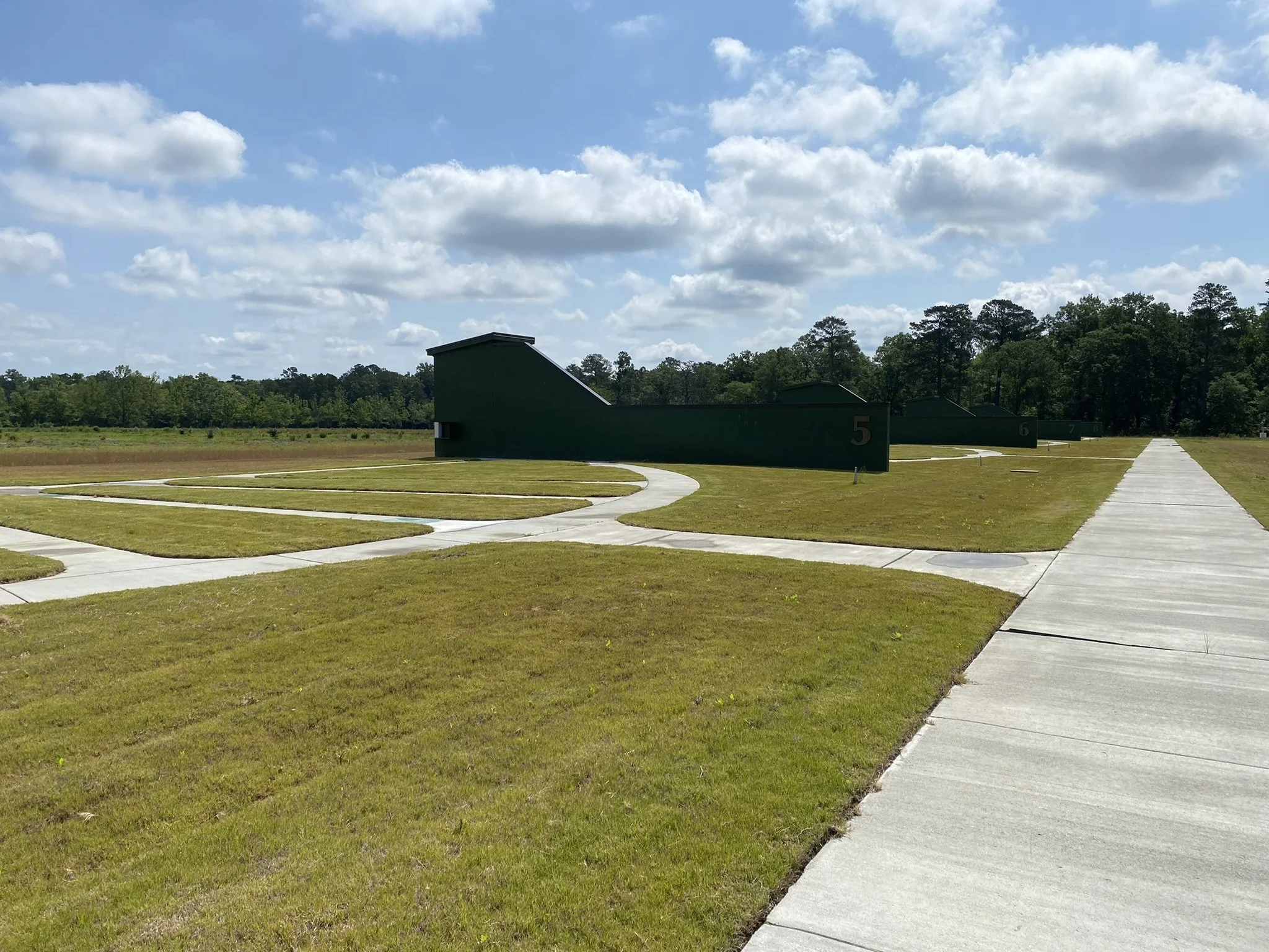 A shooting range with multiple targets, numbered 5 to 8, situated along a concrete path in a grassy field. There are trees and a blue sky with some clouds in the background.