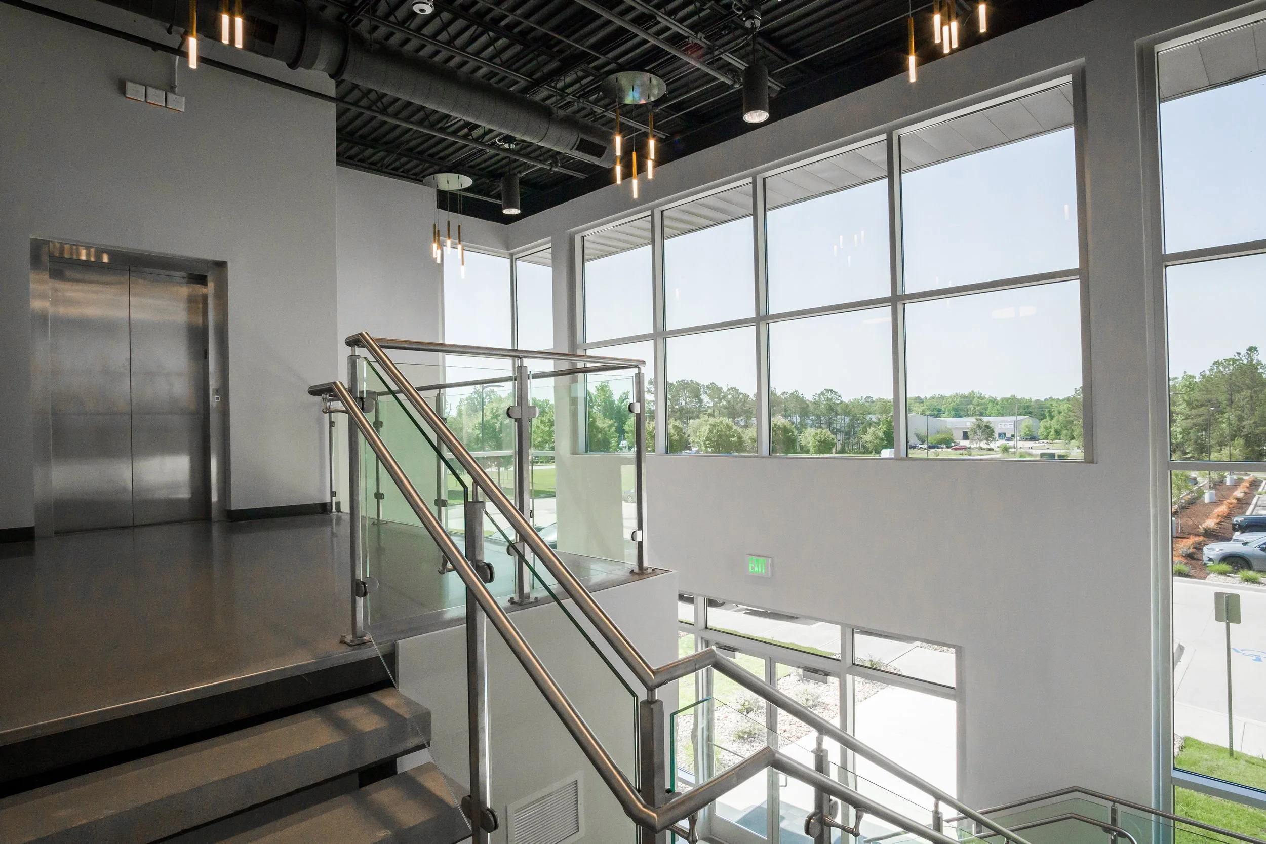 Interior view of a modern building with large glass windows, a staircase with metal railing, and an elevator. The ceiling has exposed ductwork and hanging lights. Outside, a parking lot and greenery are visible.