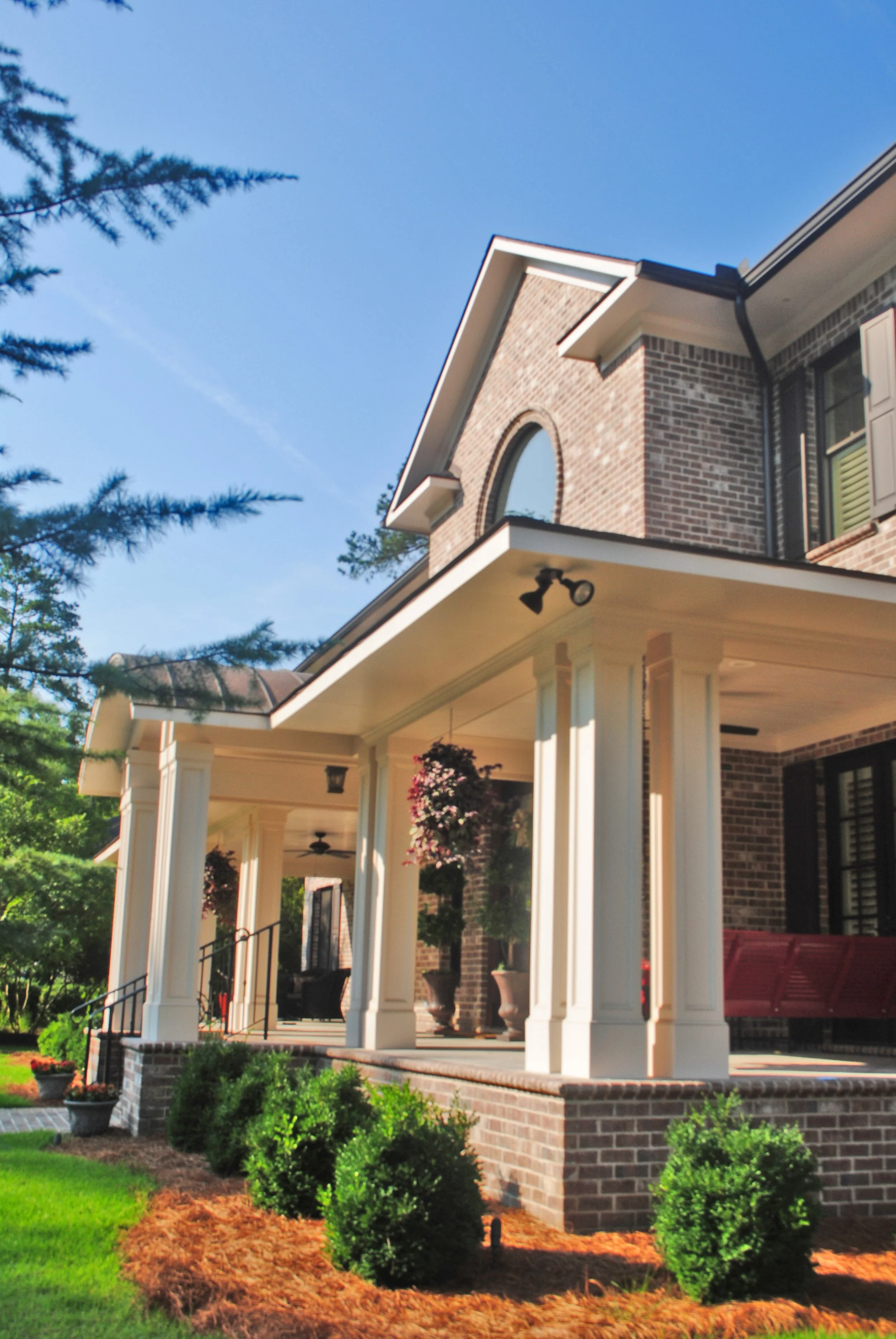 Front porch of a brick house with white columns, hanging flower baskets, and landscaped bushes, with a blue sky background.
