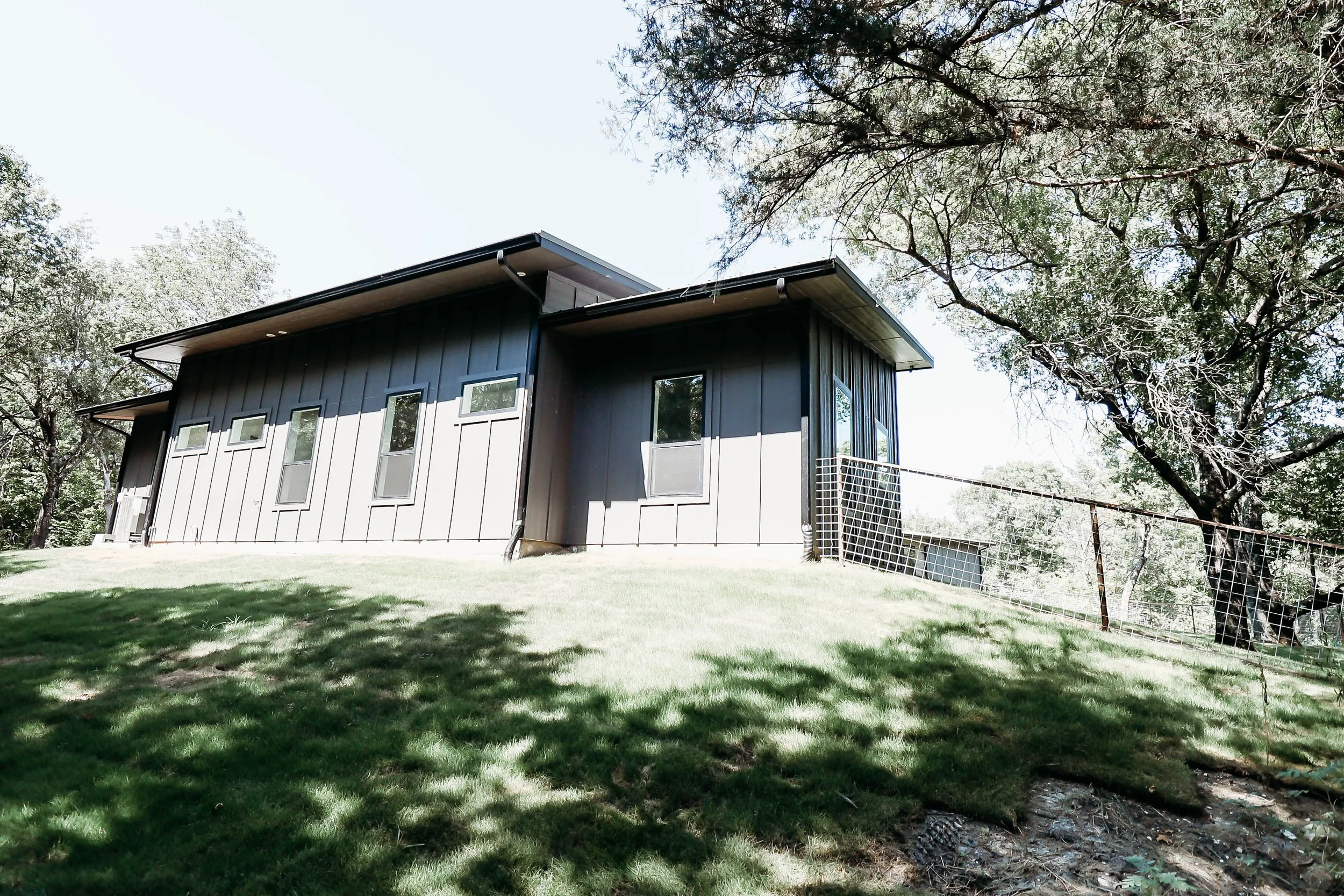Modern two-story house with black exterior, located outdoors among trees with a grassy yard.