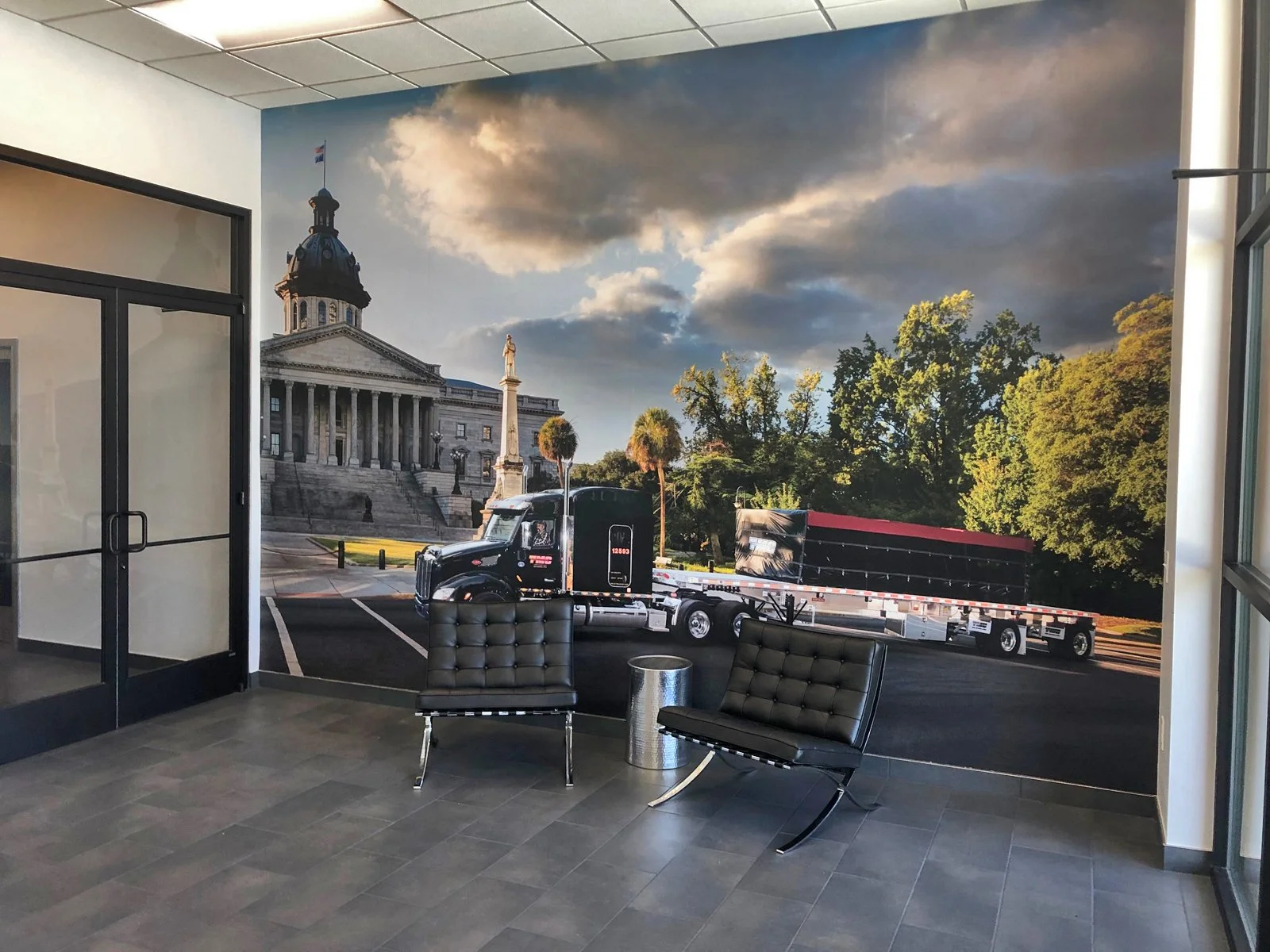 Interior of a room with a mural wall depicting a cityscape with a historic building and trees, with two black chairs and a small round silver table.