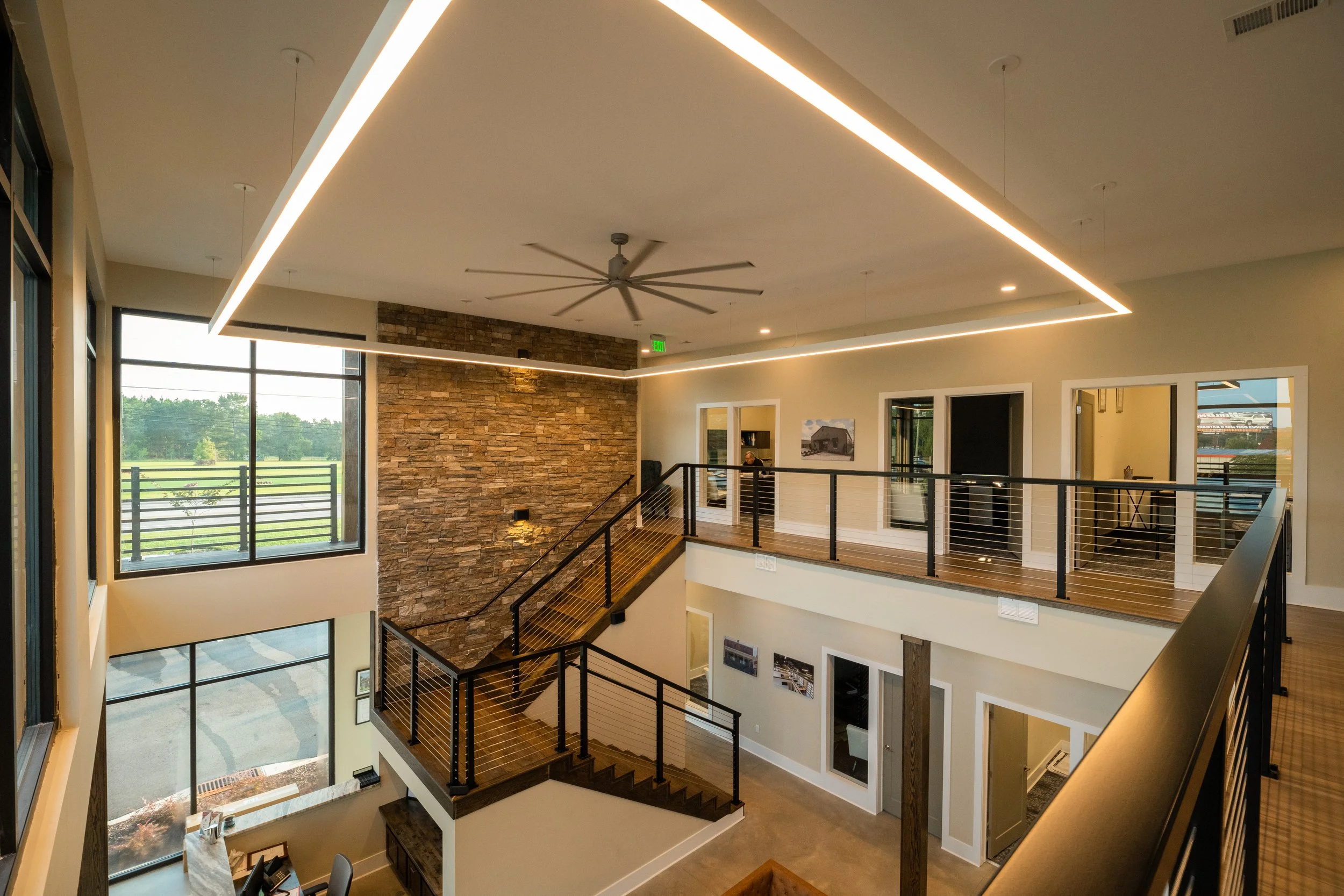 Modern open two-story interior with large windows, a brick accent wall, a ceiling fan, and linear ceiling lights.