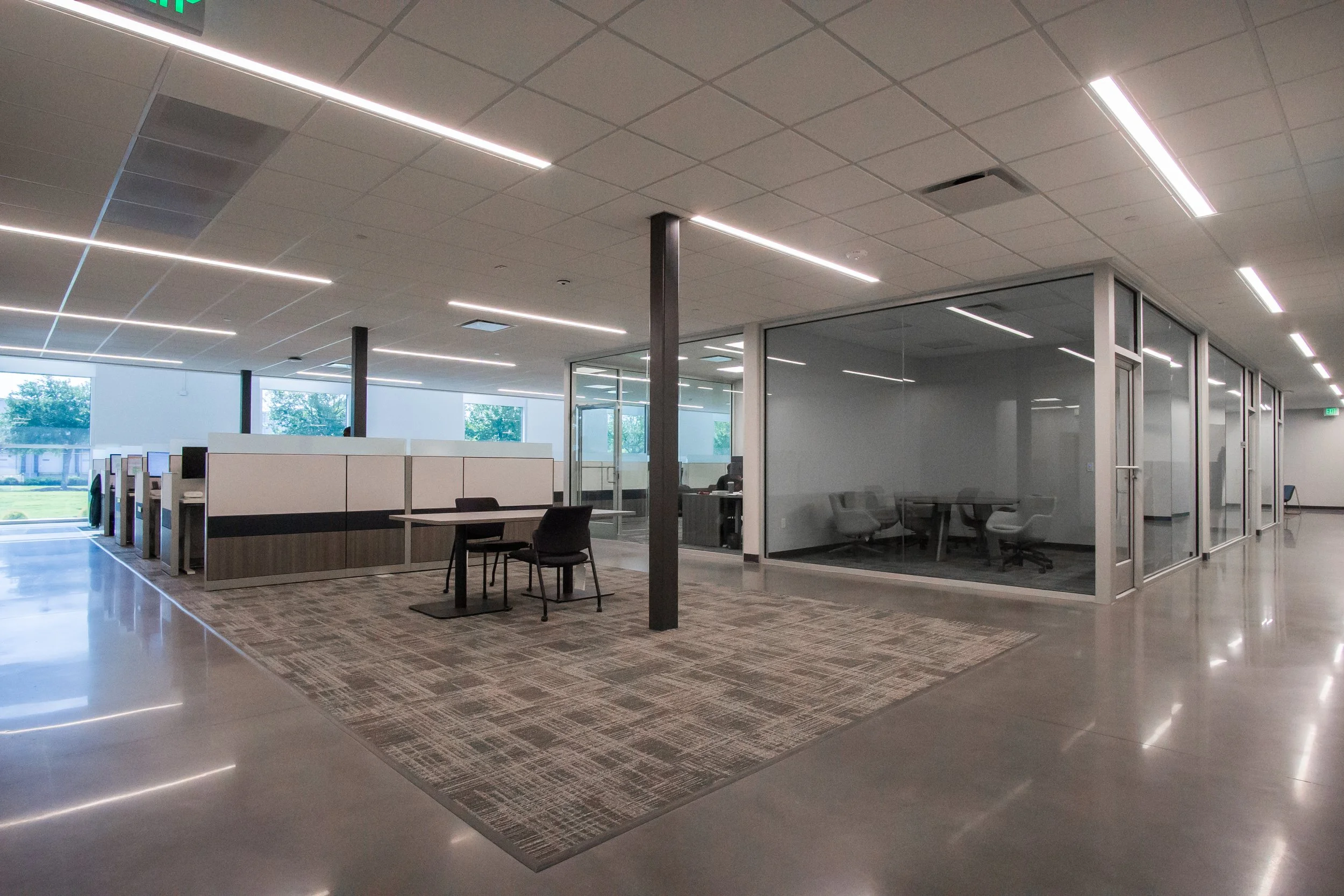 Empty office space with glass conference rooms, chairs, desks, large windows, ceiling lights, and a carpeted area.