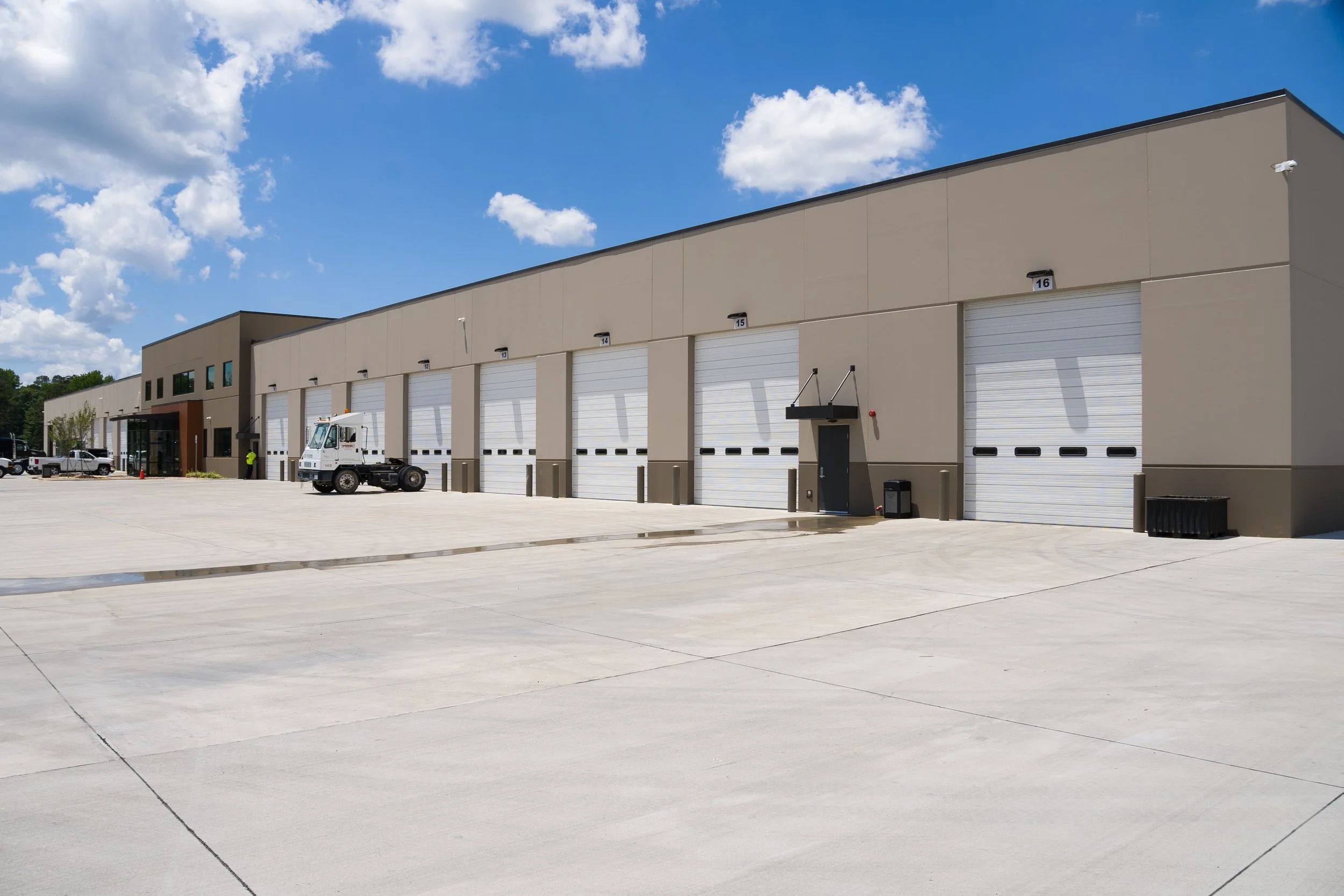 An industrial building with multiple large garage doors, a parking lot, and a truck in front under a partly cloudy sky.