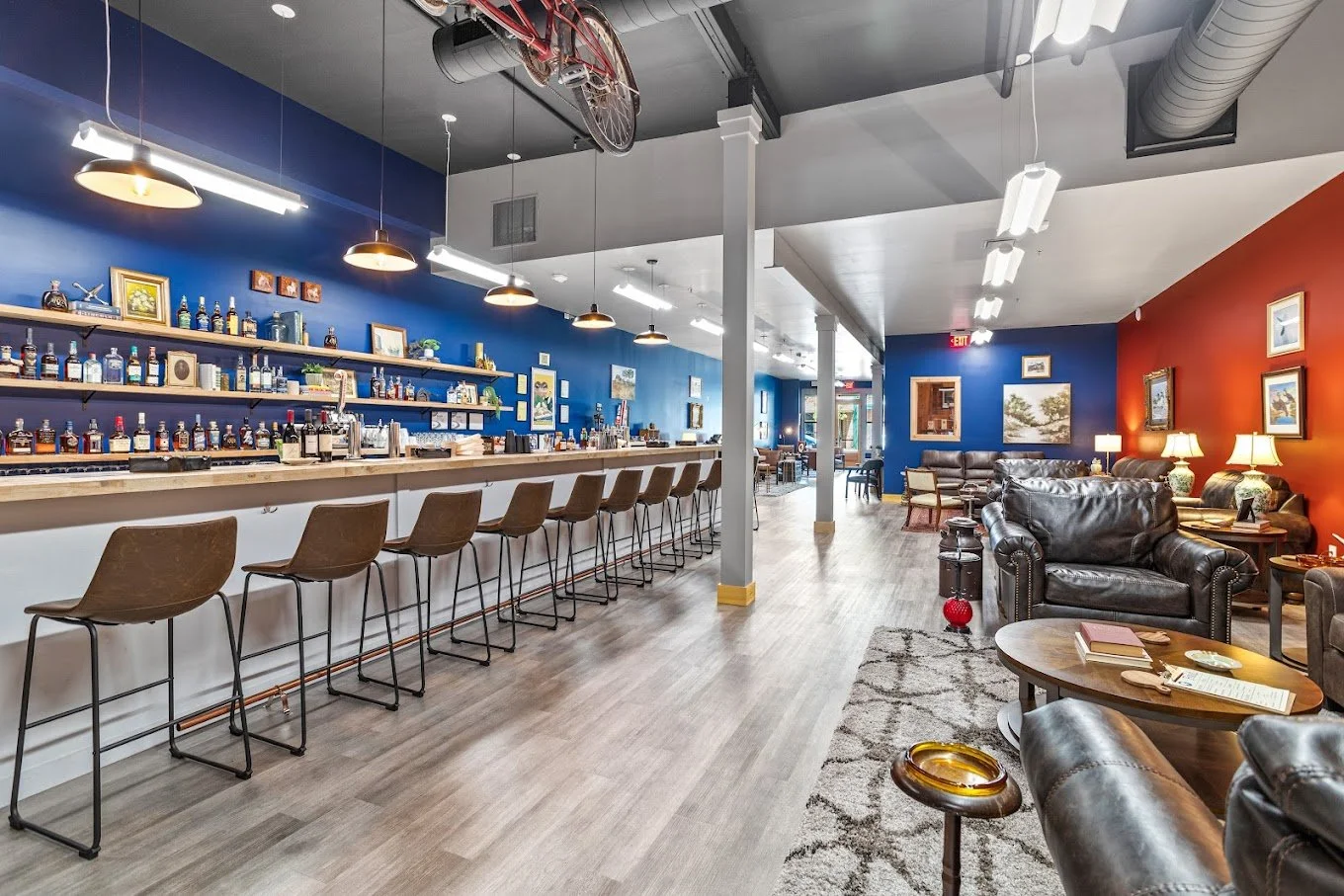 Interior of a cozy bar and lounge area with a long bar counter on the left with bar stools, colorful walls, various seating options including couches and chairs, and decorative artwork on the walls.