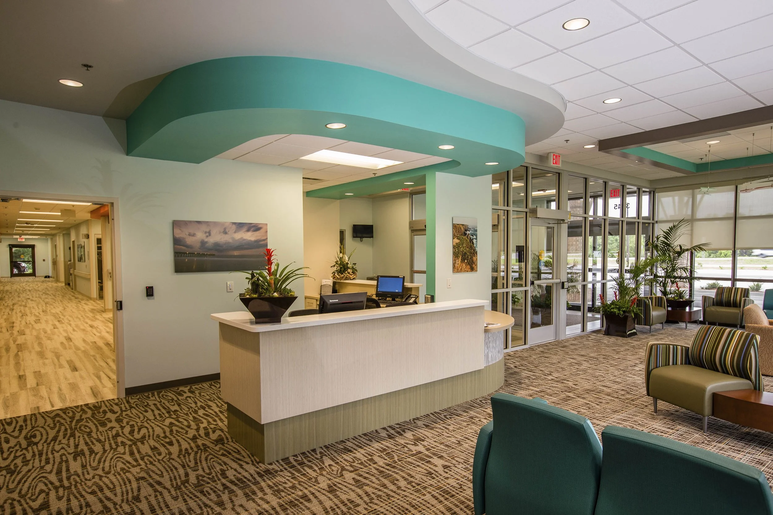Hospital or clinic lobby with waiting area, reception desk, potted plants, large windows, and artwork on the walls.