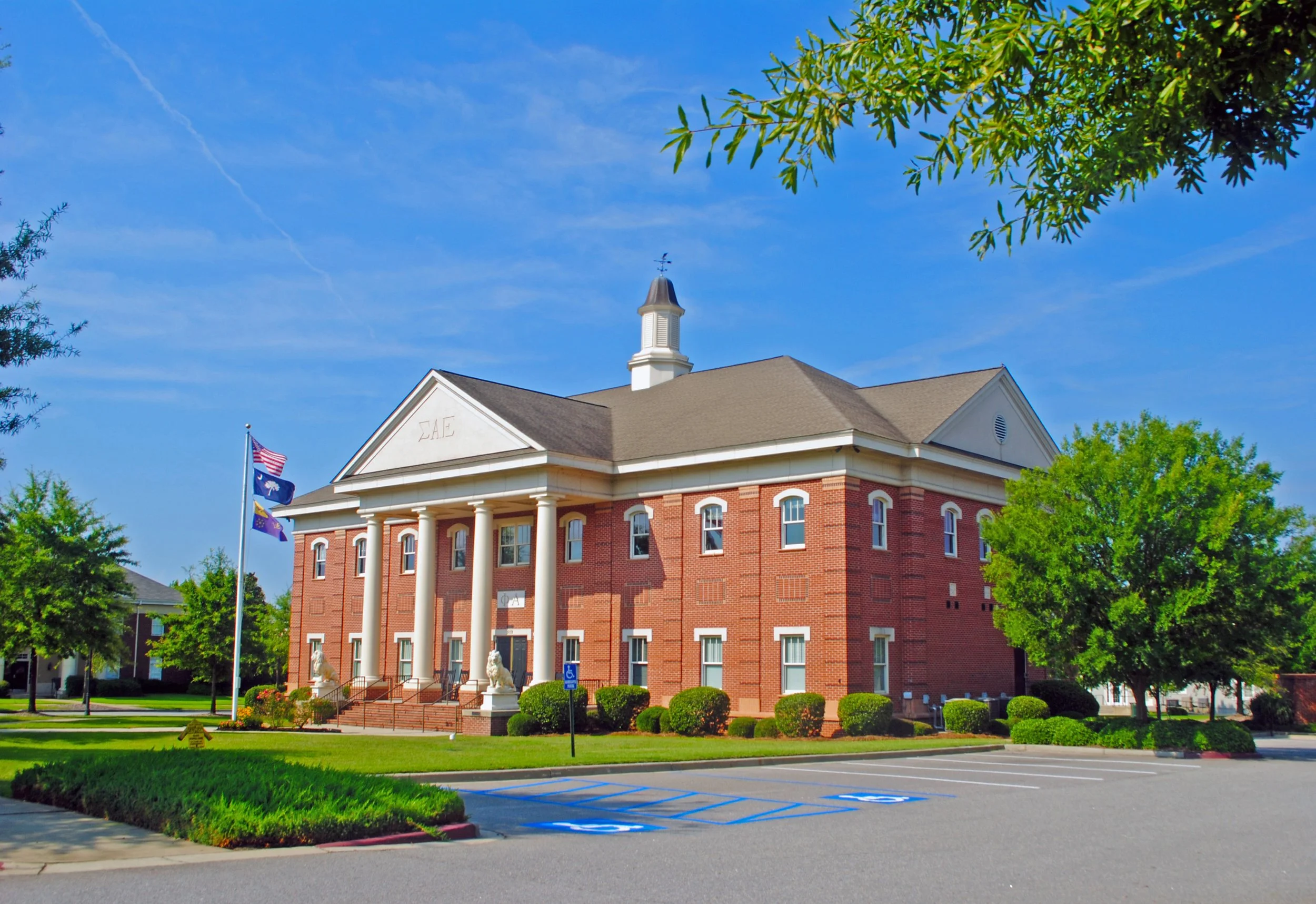 A red-brick building with a white clock tower and columns, surrounded by green trees, with parking spaces in the foreground and blue sky above.