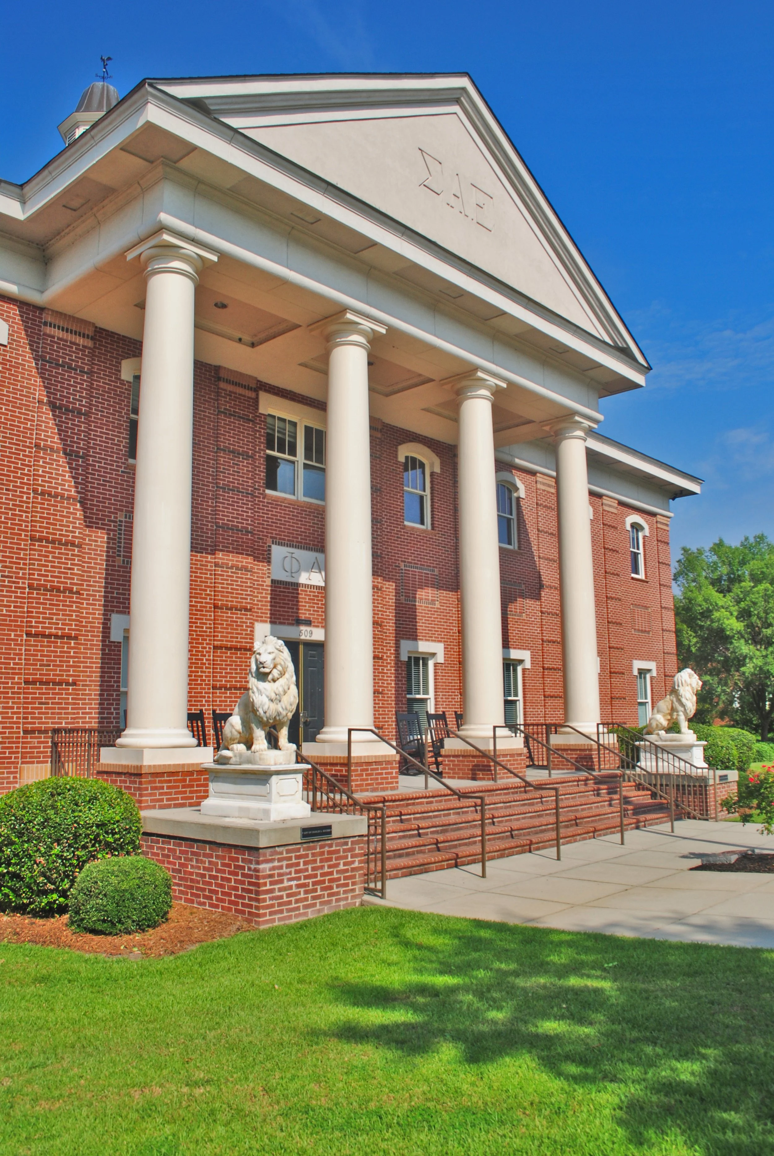 A brick fraternity house with four large white columns at the entrance, topped with lion statues on pedestals. The house has a triangular pediment with Greek letters 'ΦΔΘ' and an American flag weathervane on the roof. There are stairs with metal railings leading to the door and a well-maintained green lawn in front.