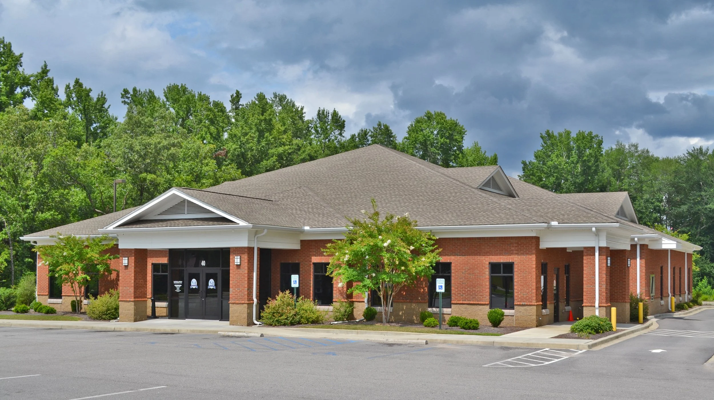 A brick building with a pitched roof and glass doors, surrounded by small trees and landscaped bushes. There are two parking spaces in front, one marked for handicapped parking.