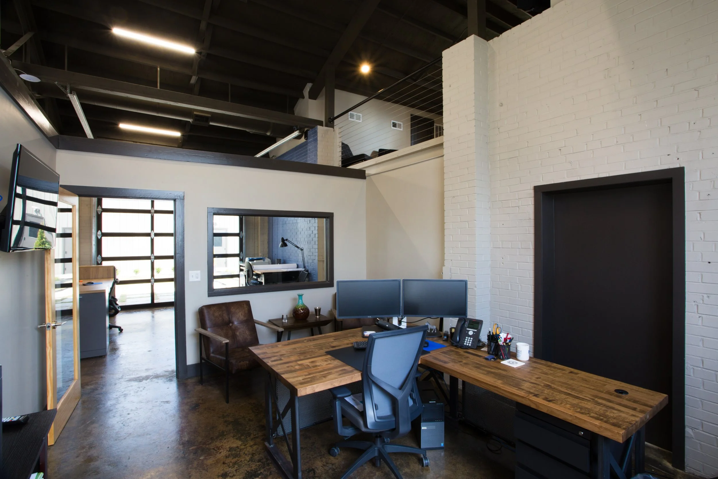 Office space with a large wooden desk, multiple monitors, office supplies, and a brown leather chair under a white brick wall. There's a windowed door leading to another room with a desk and lamp. An open loft with railing is visible above, and the c