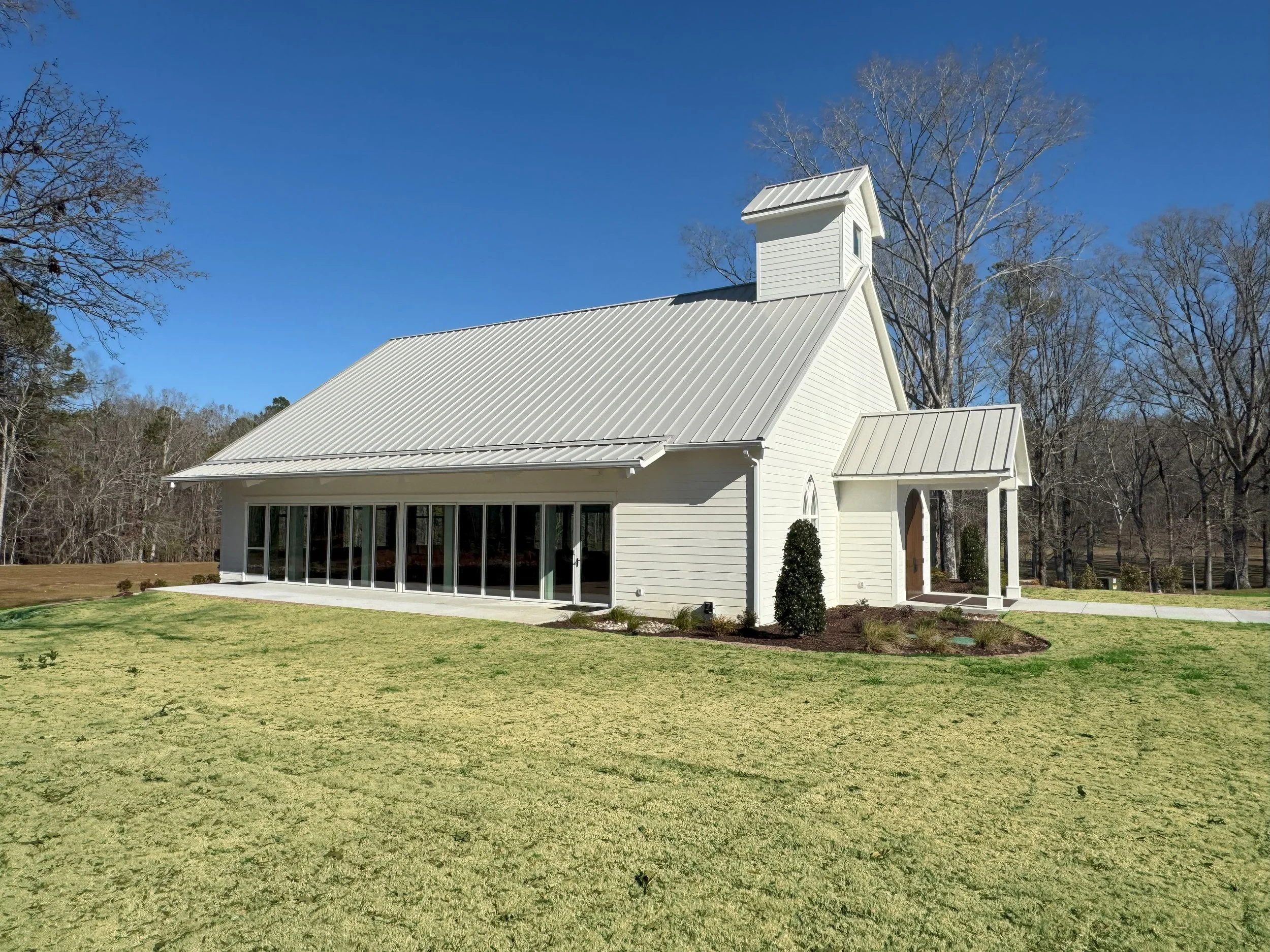 White church with a steep metallic roof, a small tower, large windows, and a wooden door, surrounded by trees with bare branches and green grass, under a clear blue sky.