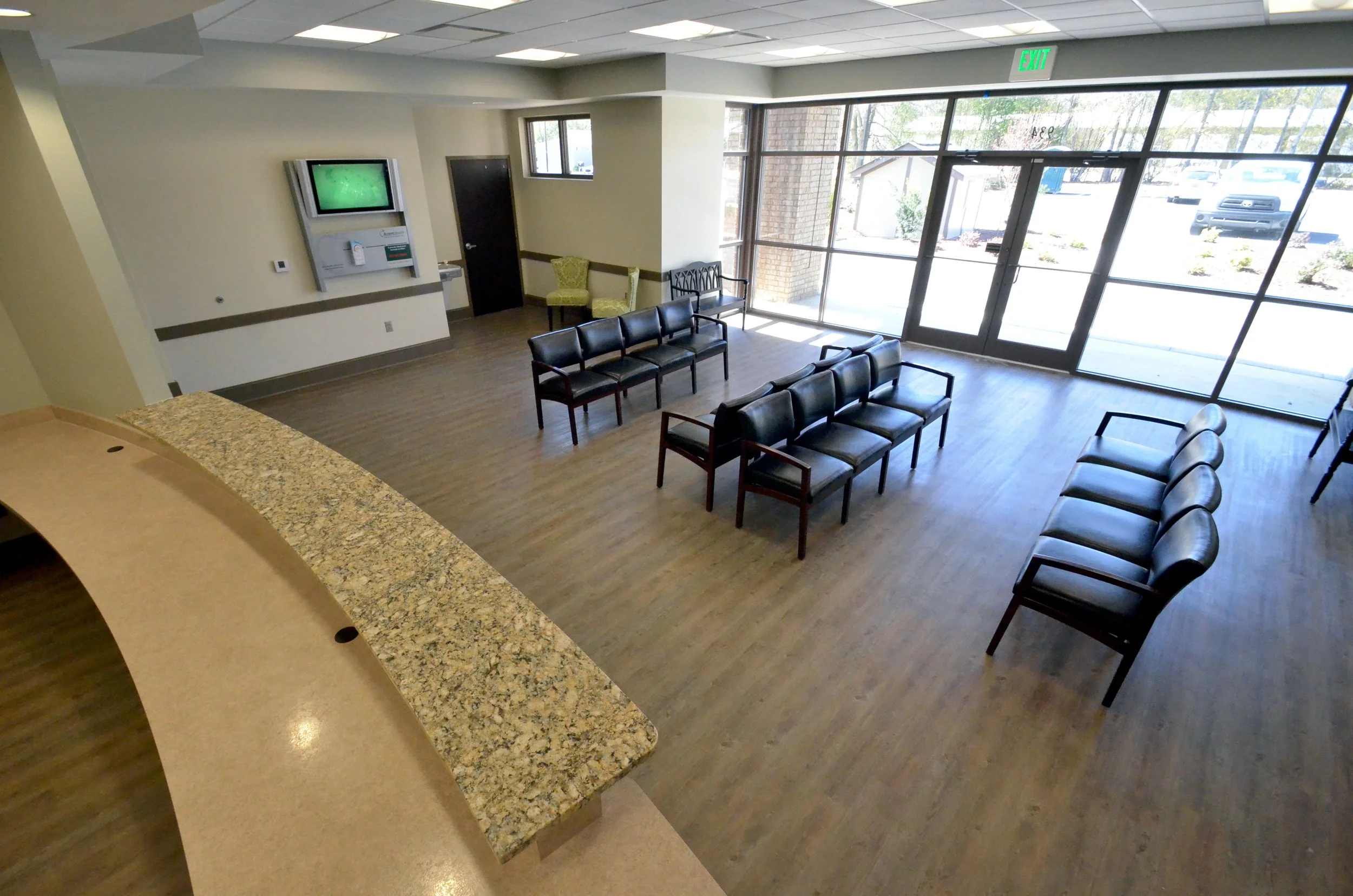Empty waiting area with black chairs, two green chairs, and a large glass front door and window showing outside parking lot.
