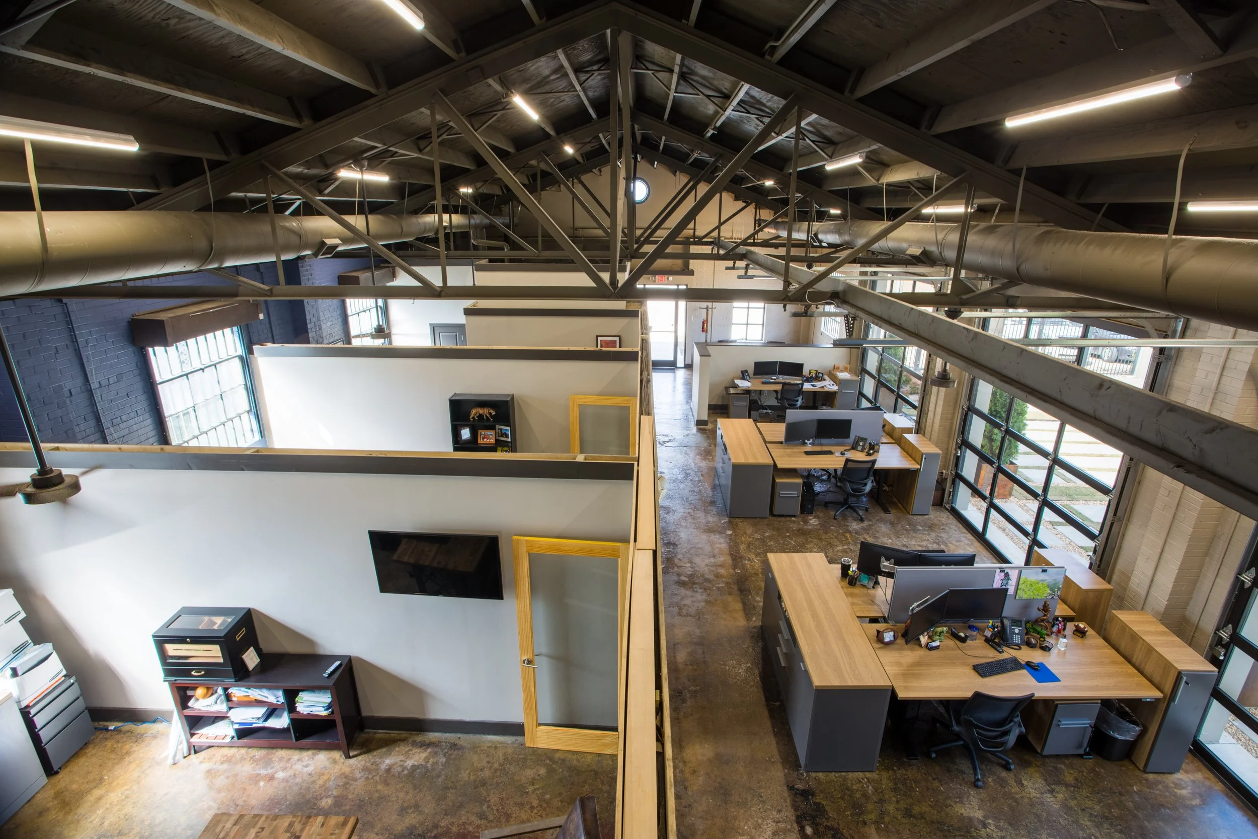 An office space with multiple workstations, computers, and office supplies on desks. Large windows allow natural light to enter, and there are visible ductwork and metal beams on the ceiling, giving the space an industrial look.