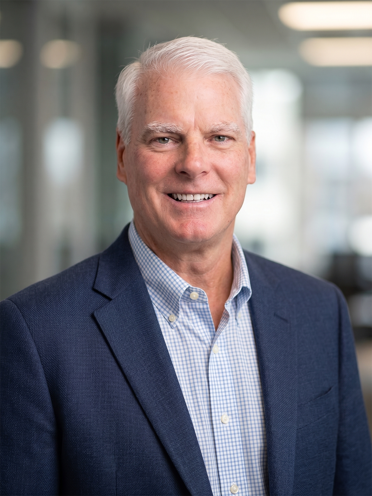 Headshot of a smiling middle-aged man with white hair, dressed in a blue blazer and a light blue checkered shirt, in a professional indoor setting.