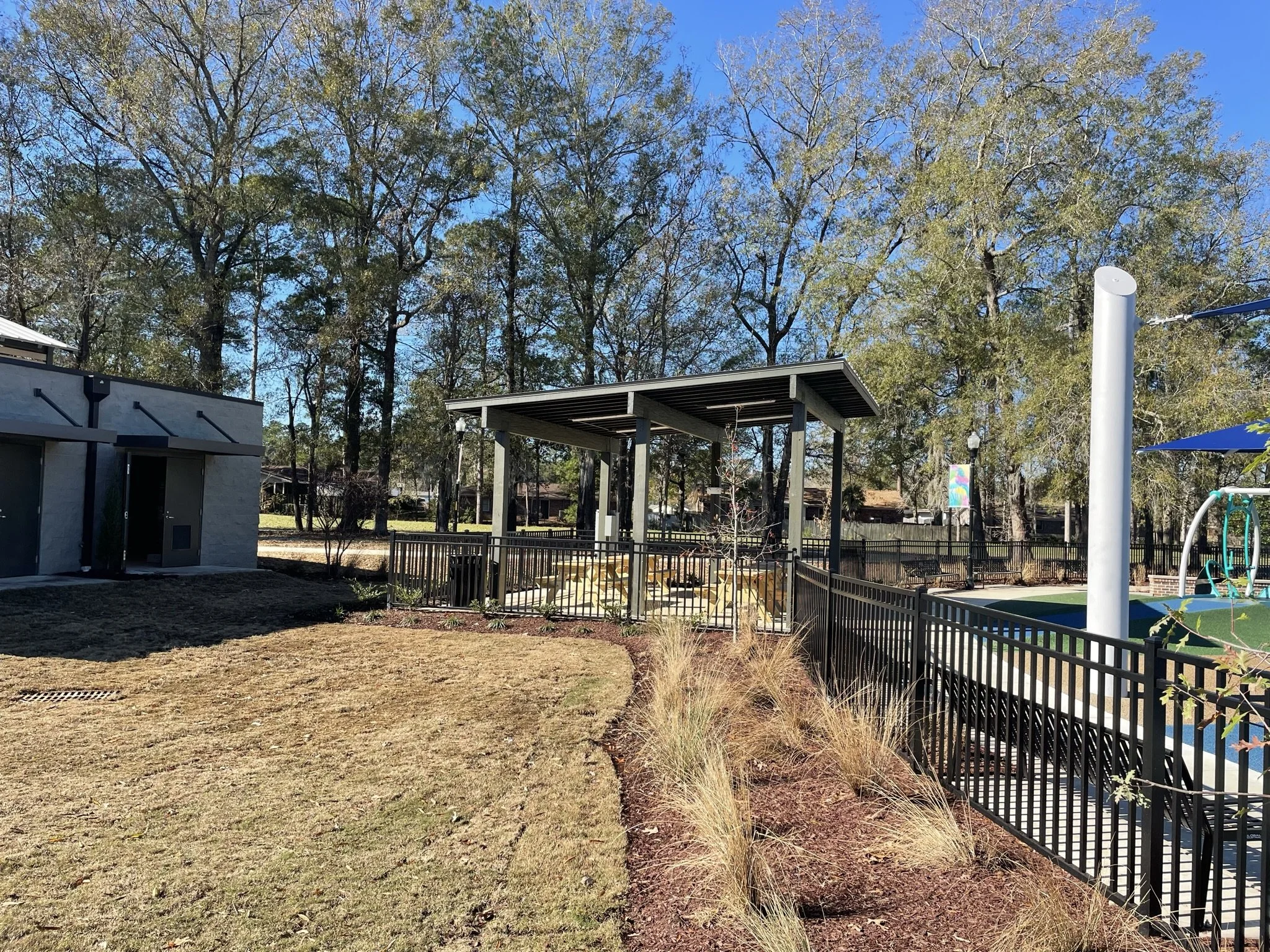 An outdoor area with a lawn, a fenced playground, and a structure with a roof. There are trees with sparse leaves and a clear blue sky.