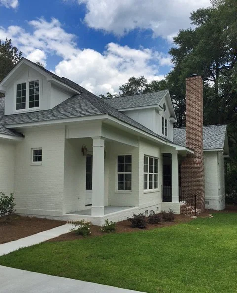 White two-story house with a brick chimney, front porch, and multiple windows, surrounded by a green lawn and trees under a partly cloudy sky.