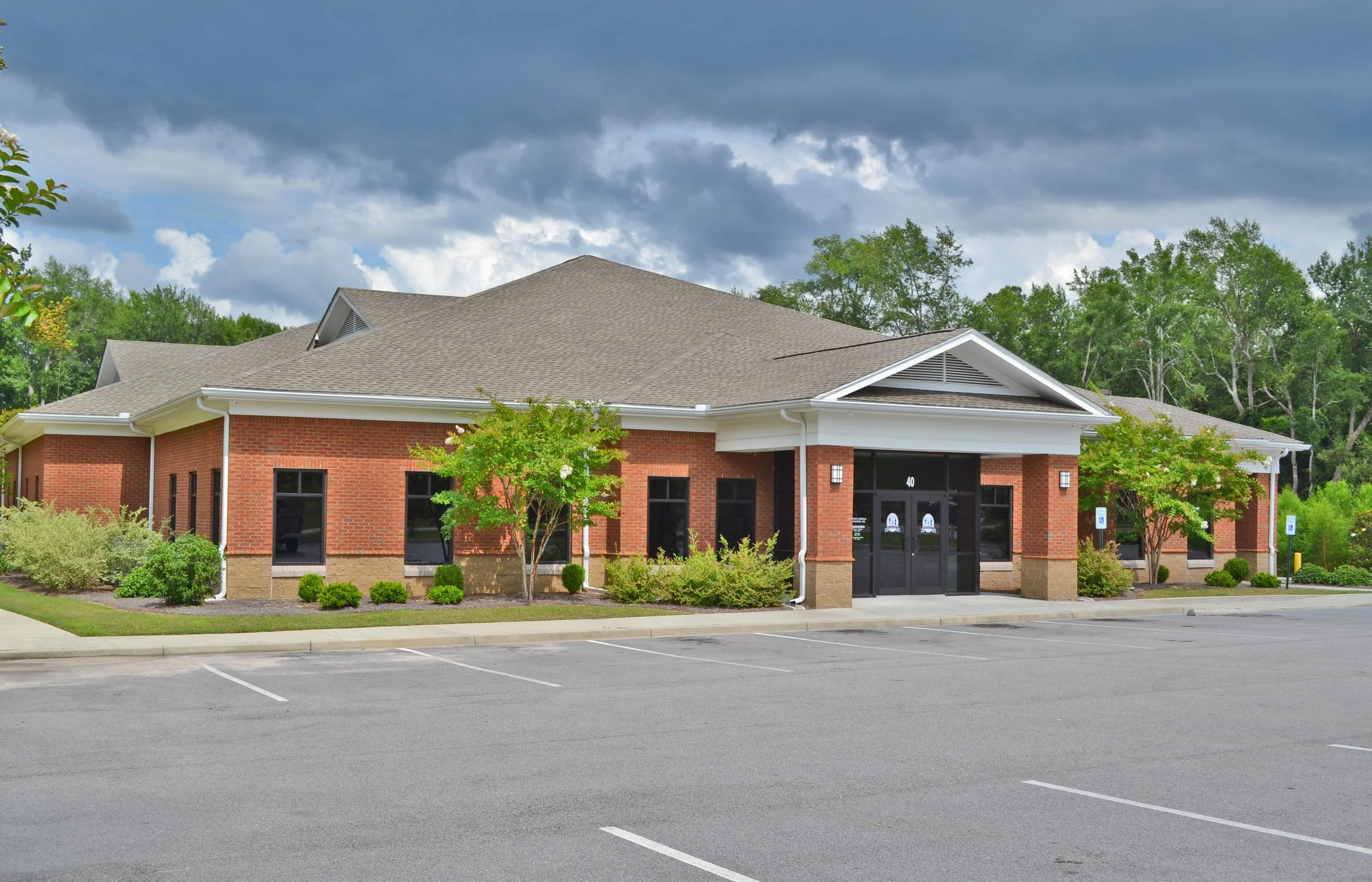 Commercial building with brick walls, large windows, and a covered entrance, surrounded by landscaping, under a cloudy sky.