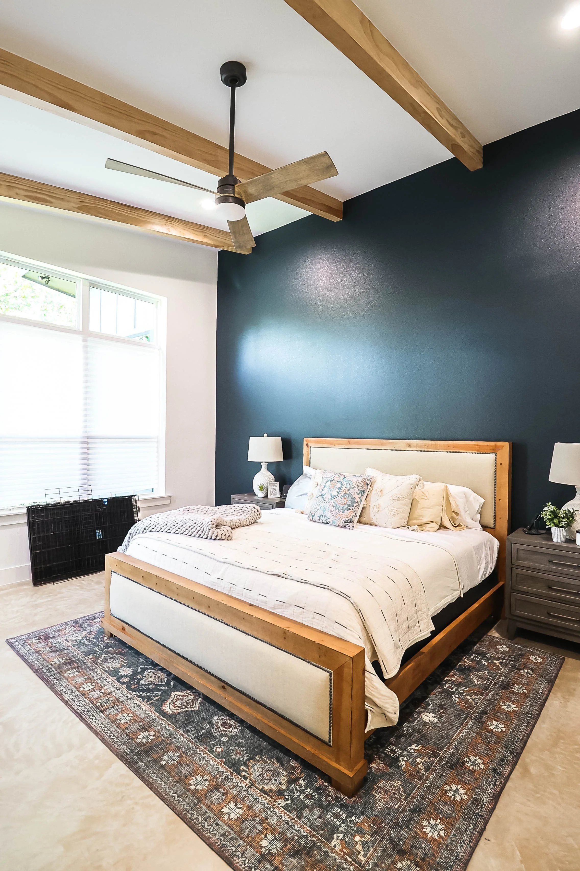 A bedroom with a dark blue accent wall, a wooden bed frame with white bedding, a patterned rug, two nightstands with lamps, and a ceiling fan with wooden blades.