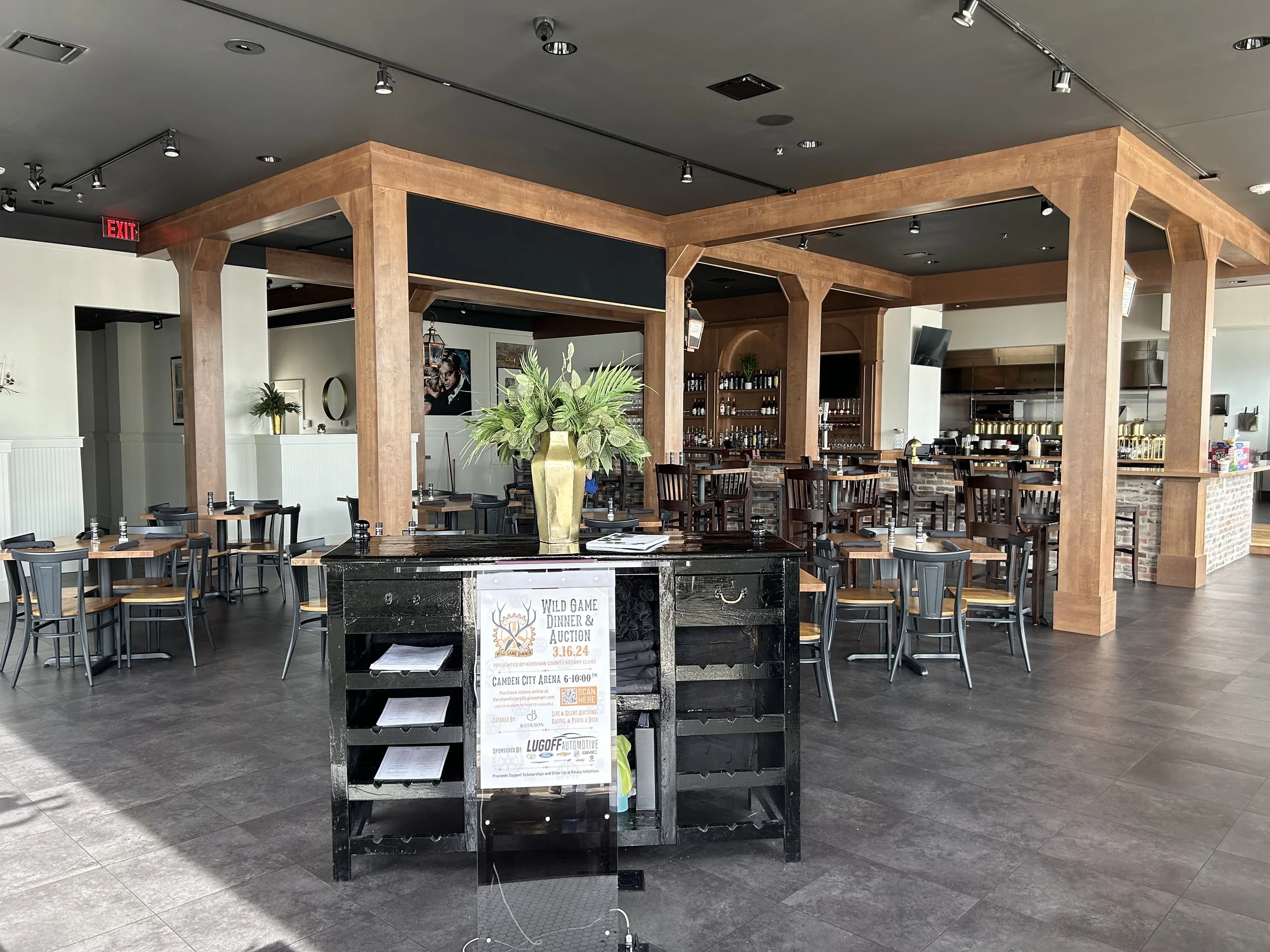 Interior of a restaurant with wooden and brick decor, seating areas, and a central black table displaying a sign for an upcoming dinner and auction event.