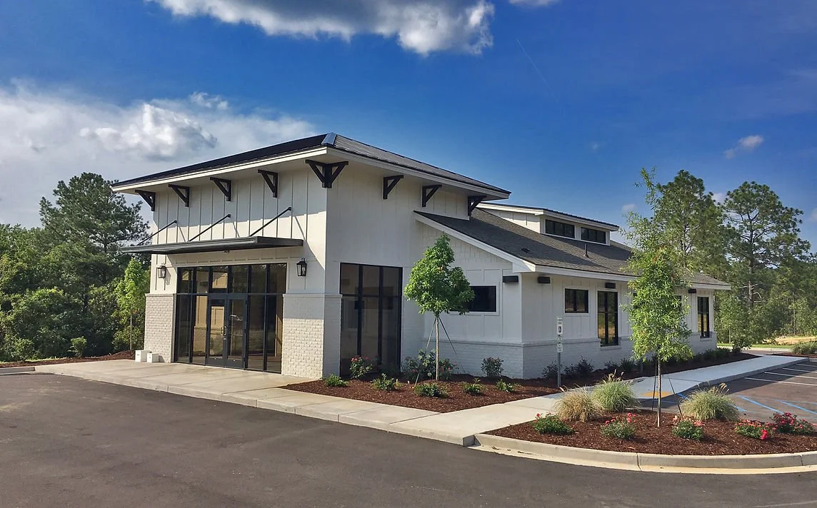 Modern white commercial building with large glass windows and landscaping, including small trees and bushes, under a partly cloudy blue sky.