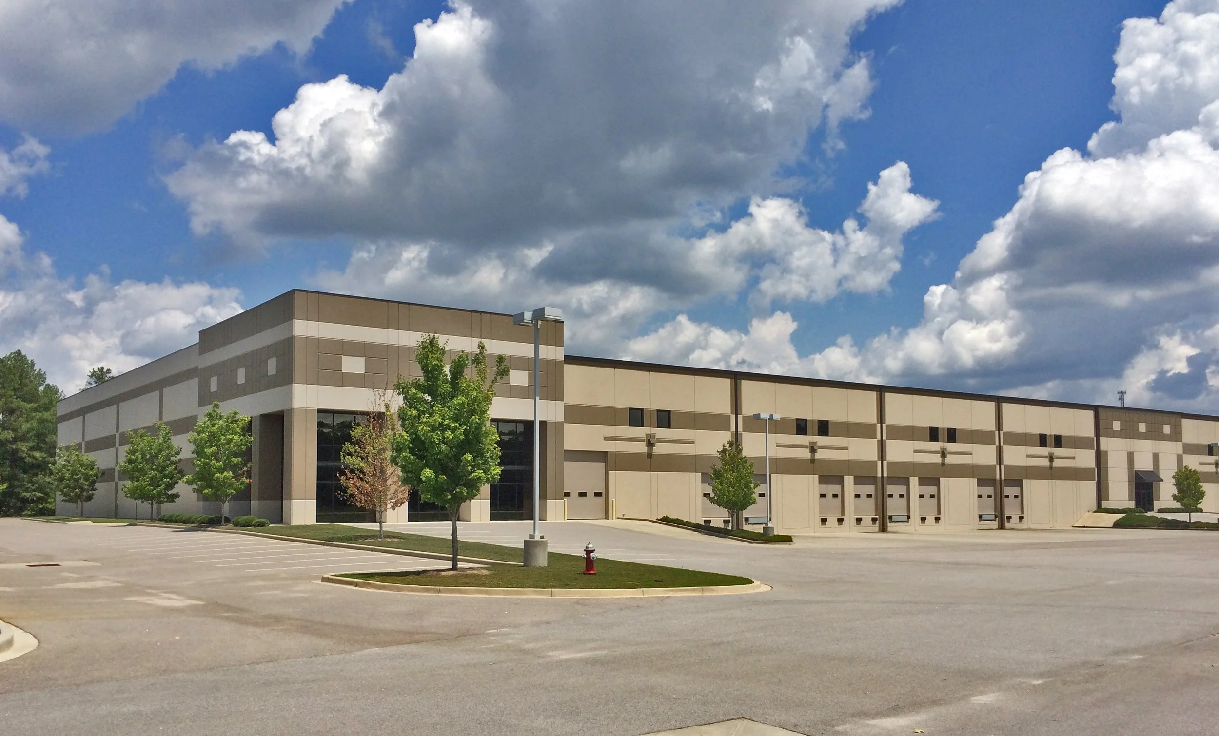 Large beige and brown commercial building with multiple garage doors in a parking lot with trees and street lamps under a partly cloudy sky.