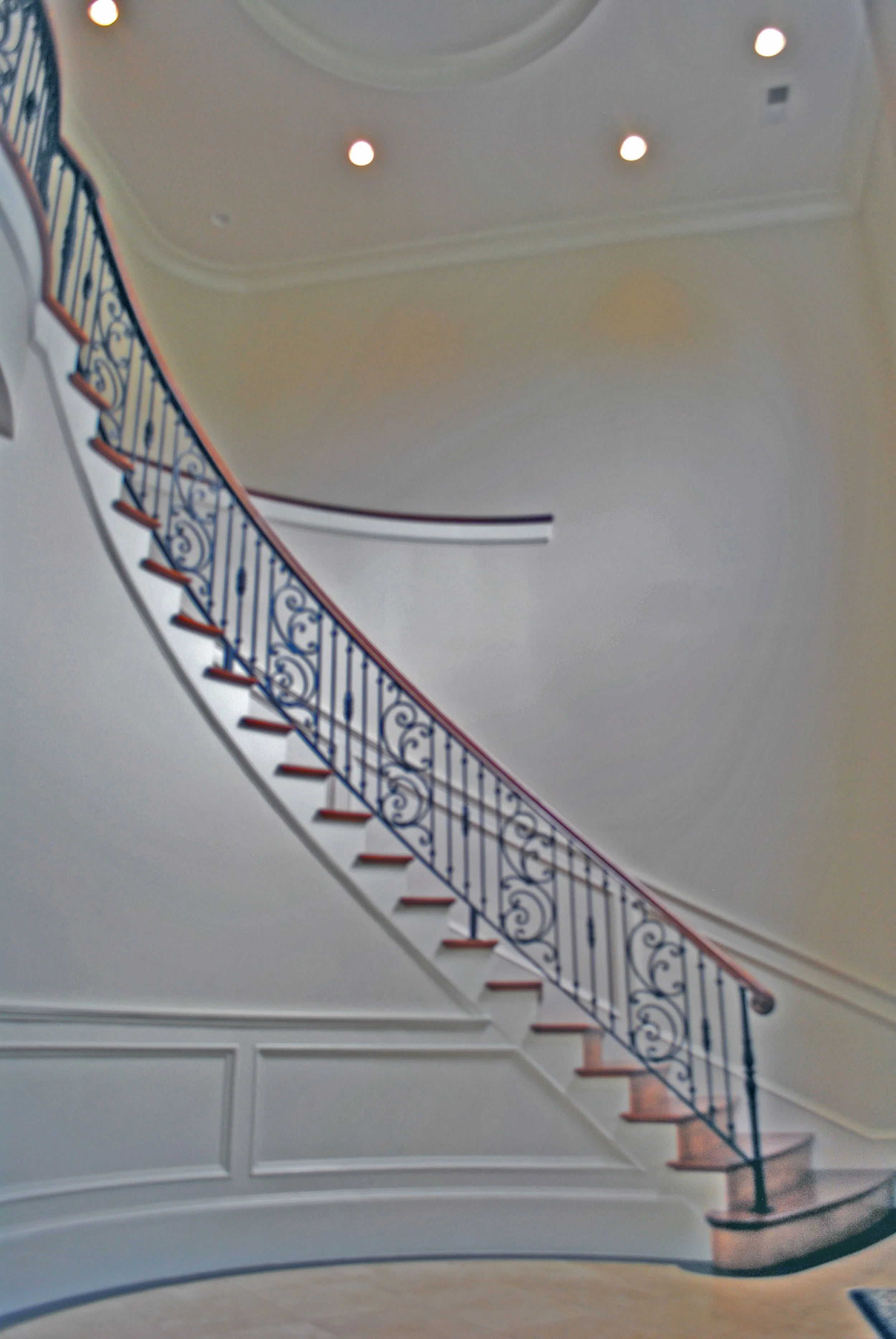 Interior view of a curved staircase with decorative iron railings, wooden steps, and a white wall with molding, ceiling lights, and a rounded ceiling detail.