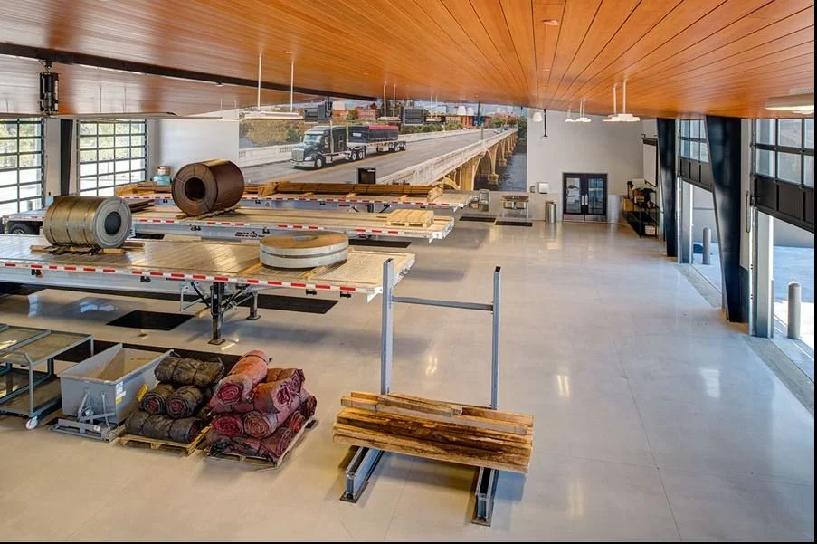 Indoor space with construction materials, including large rolls of insulation, wood planks, and carpet rolls, with a large mural of a highway scene on the back wall.