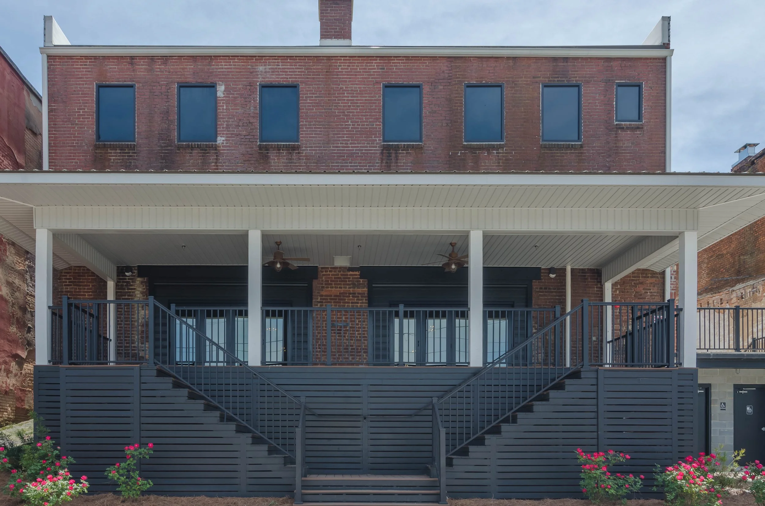 A multi-story brick building with a large porch and outdoor staircases, decorated with a black fence and colorful flowers in the front yard.
