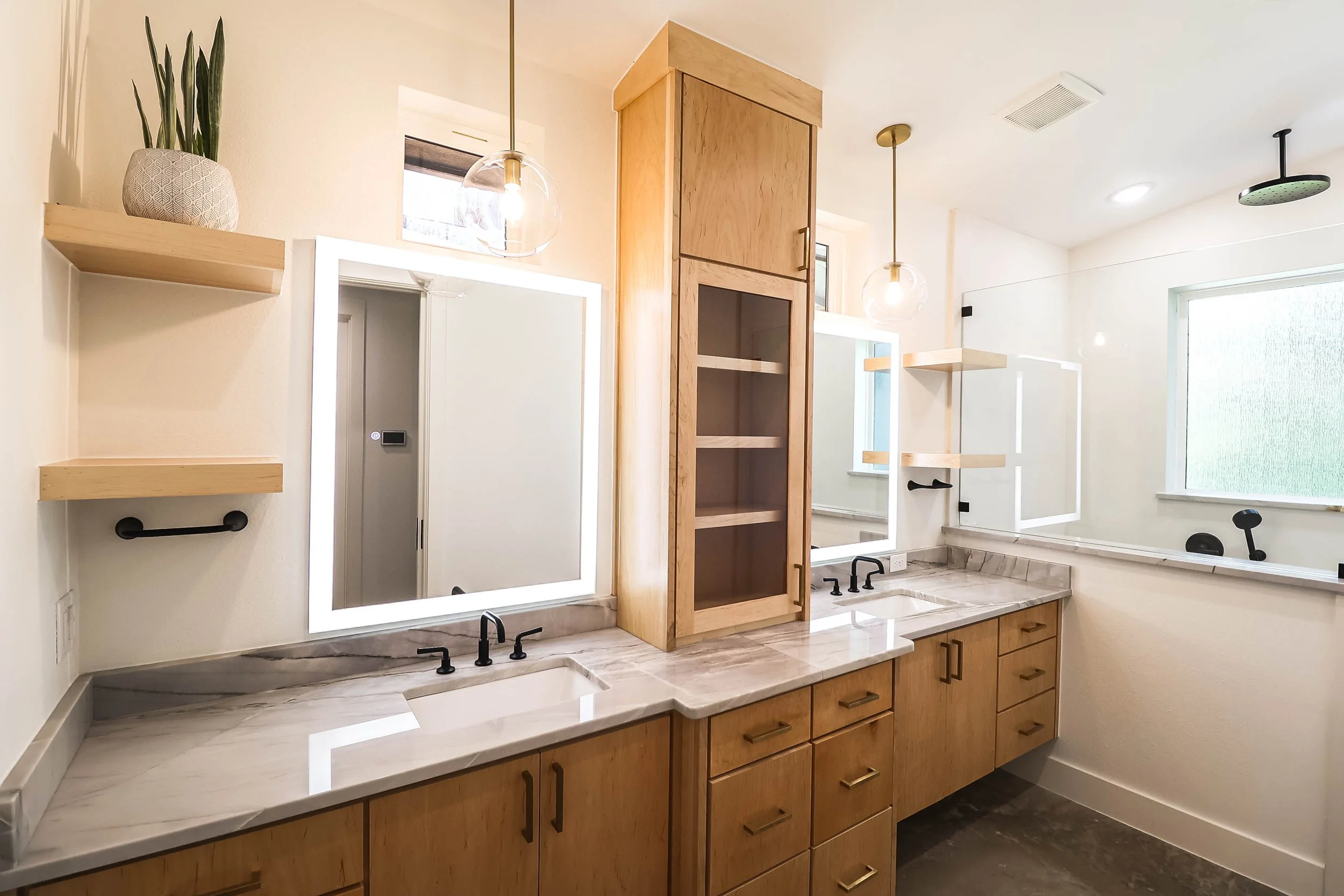 Modern bathroom with wooden cabinets, marble countertops, large mirrors with LED lighting, and black faucets. There are floating shelves, a window, and ceiling light fixtures.