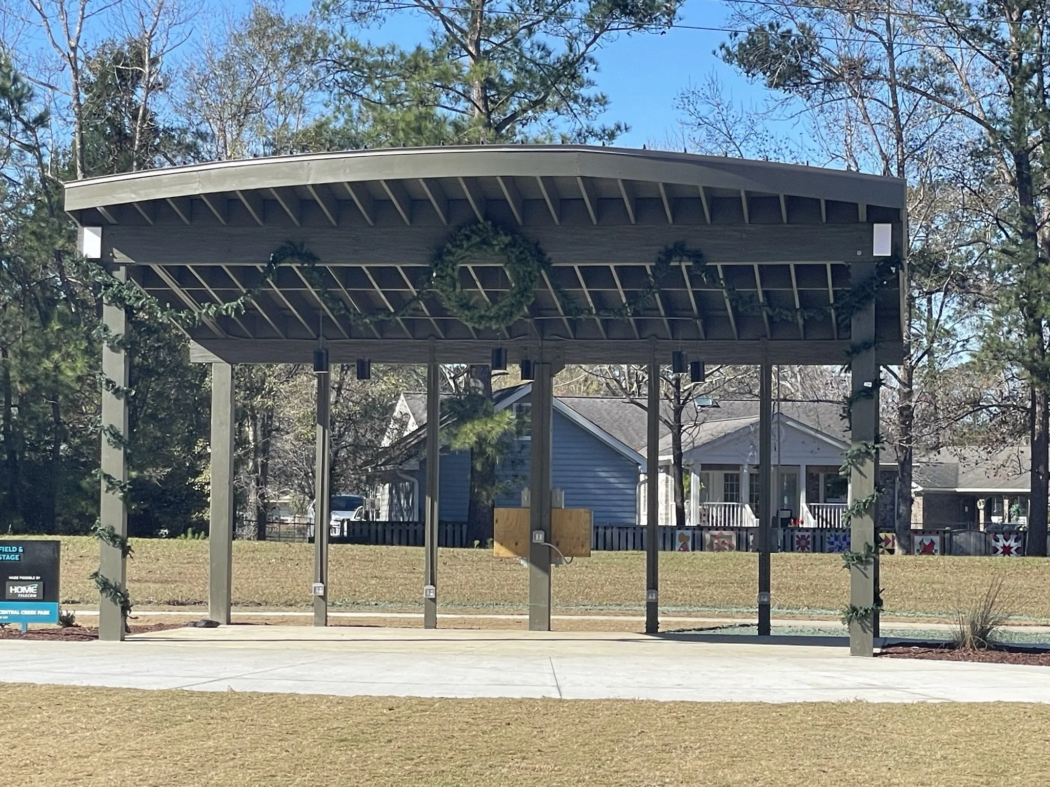 Empty outdoor stage with a roof, decorated with green wreaths, in a park area with trees and houses in the background.