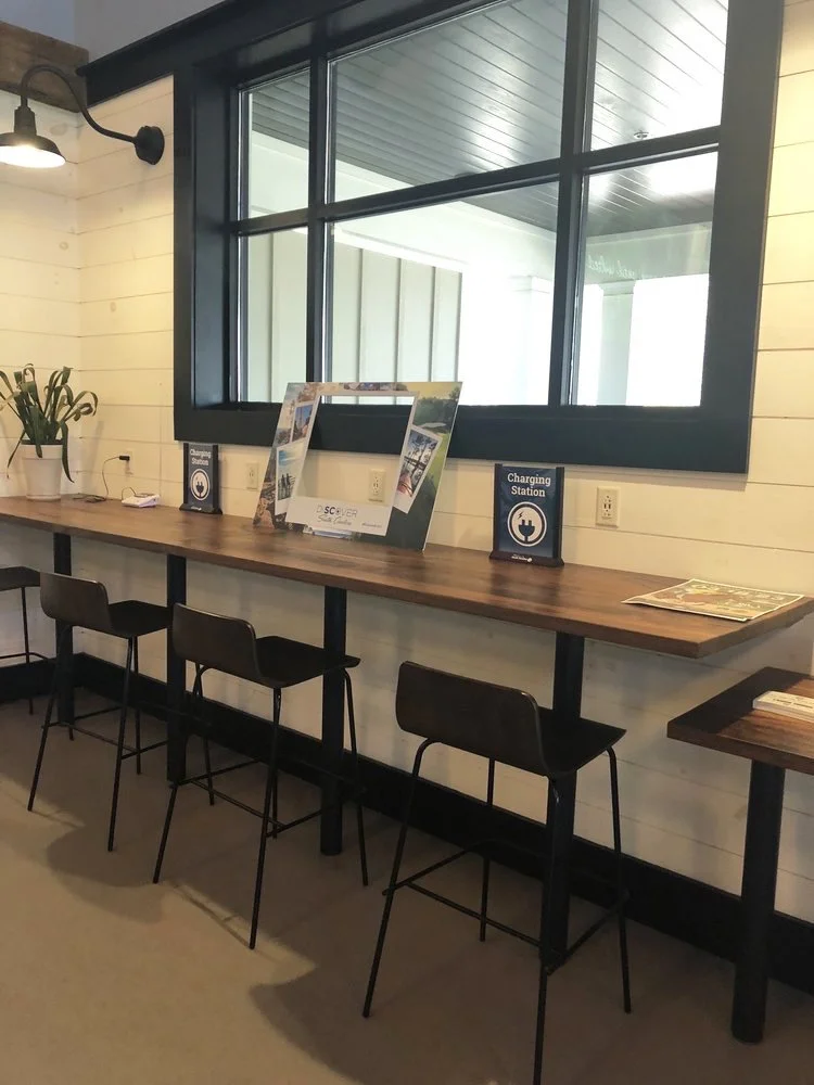 Coffee shop interior with a long wooden counter and four black chairs. There is a large window with black framing behind the counter, and on the counter are signs for a charging station and some brochures. To the left, a potted plant and wall-mounted