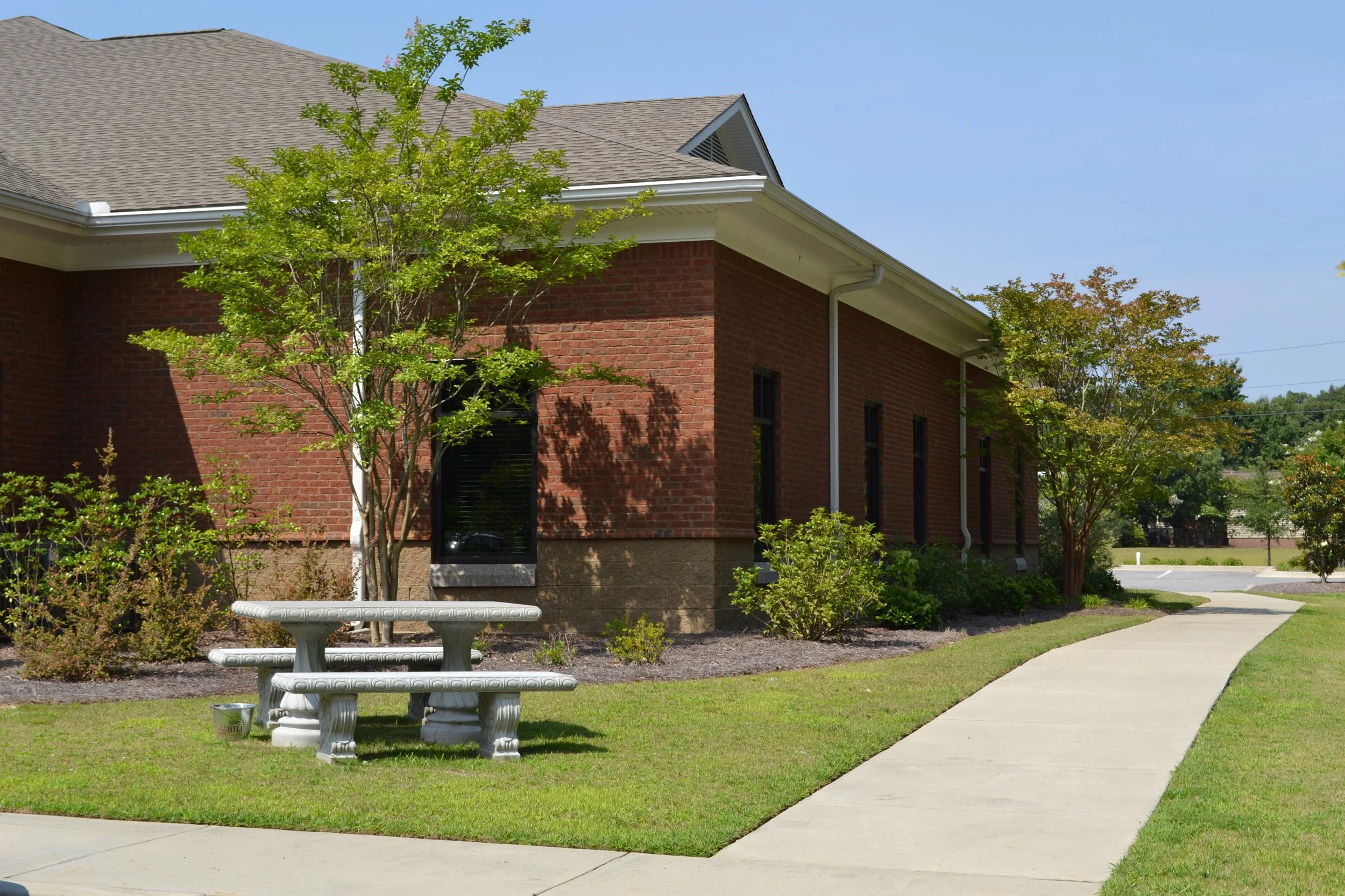 A brick building with a sloped roof, small fenced windows, and a small garden with trees and shrubs in front. There is a concrete sidewalk, a circular concrete bench, and a patch of grass.