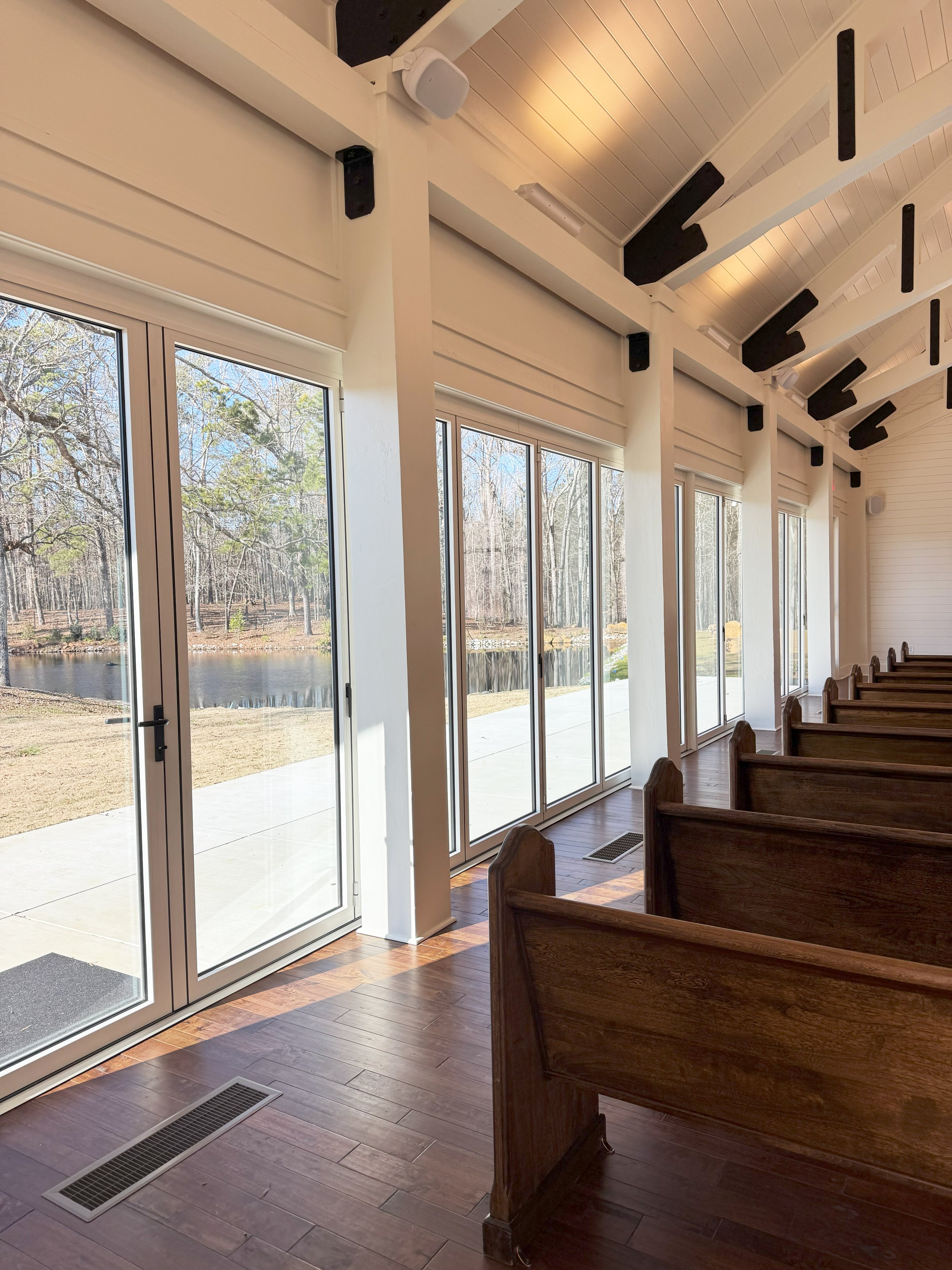 A row of large windows and glass doors overlooking a lakeside landscape with trees. Wooden pews line the interior, and sunlight streams through the windows, creating shadows on the wooden floor.