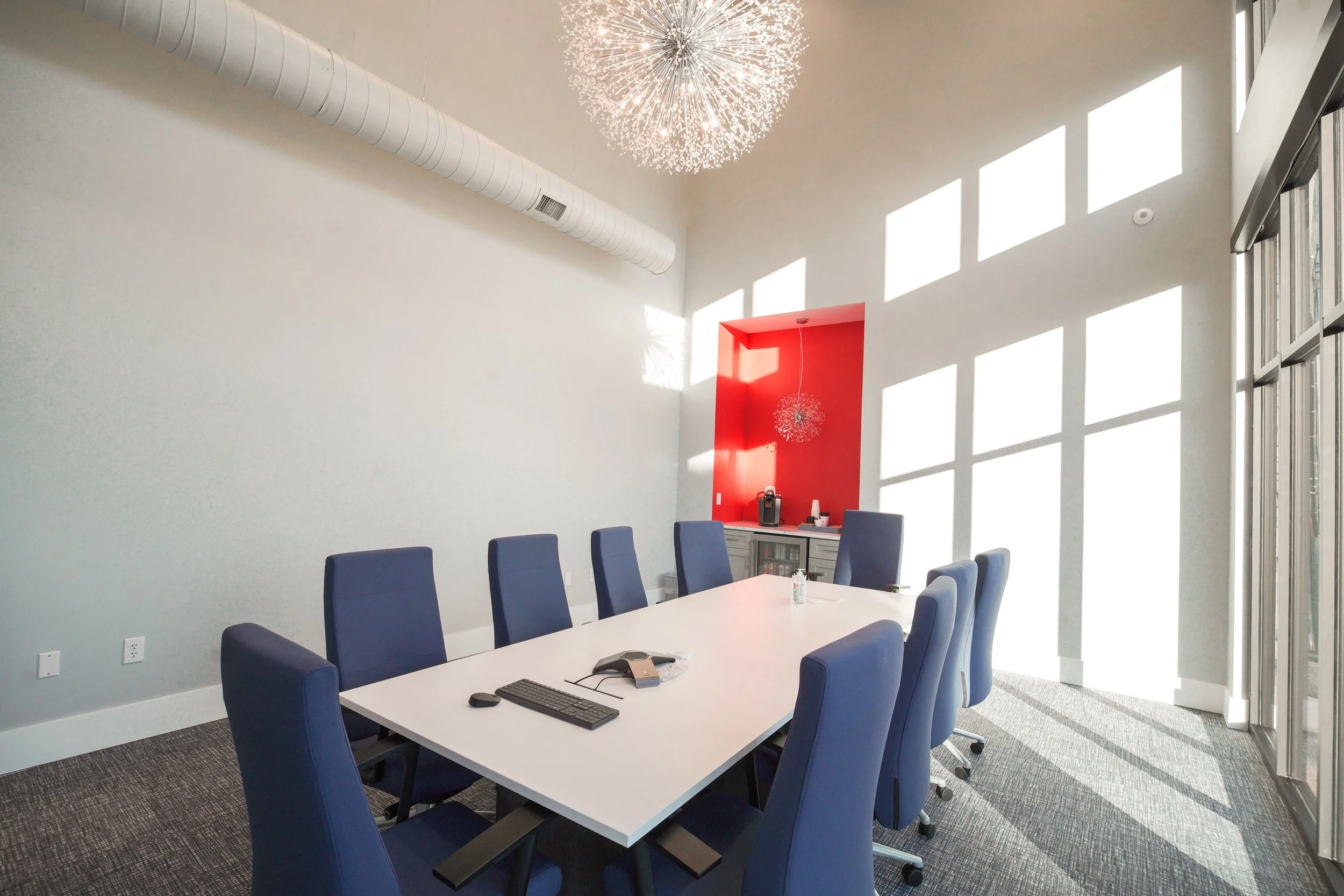 A modern conference room with a long white table surrounded by blue chairs. Natural sunlight streams through large windows, casting grid-like shadows on the white walls. The ceiling features a large, spherical chandelier and a visible white duct.