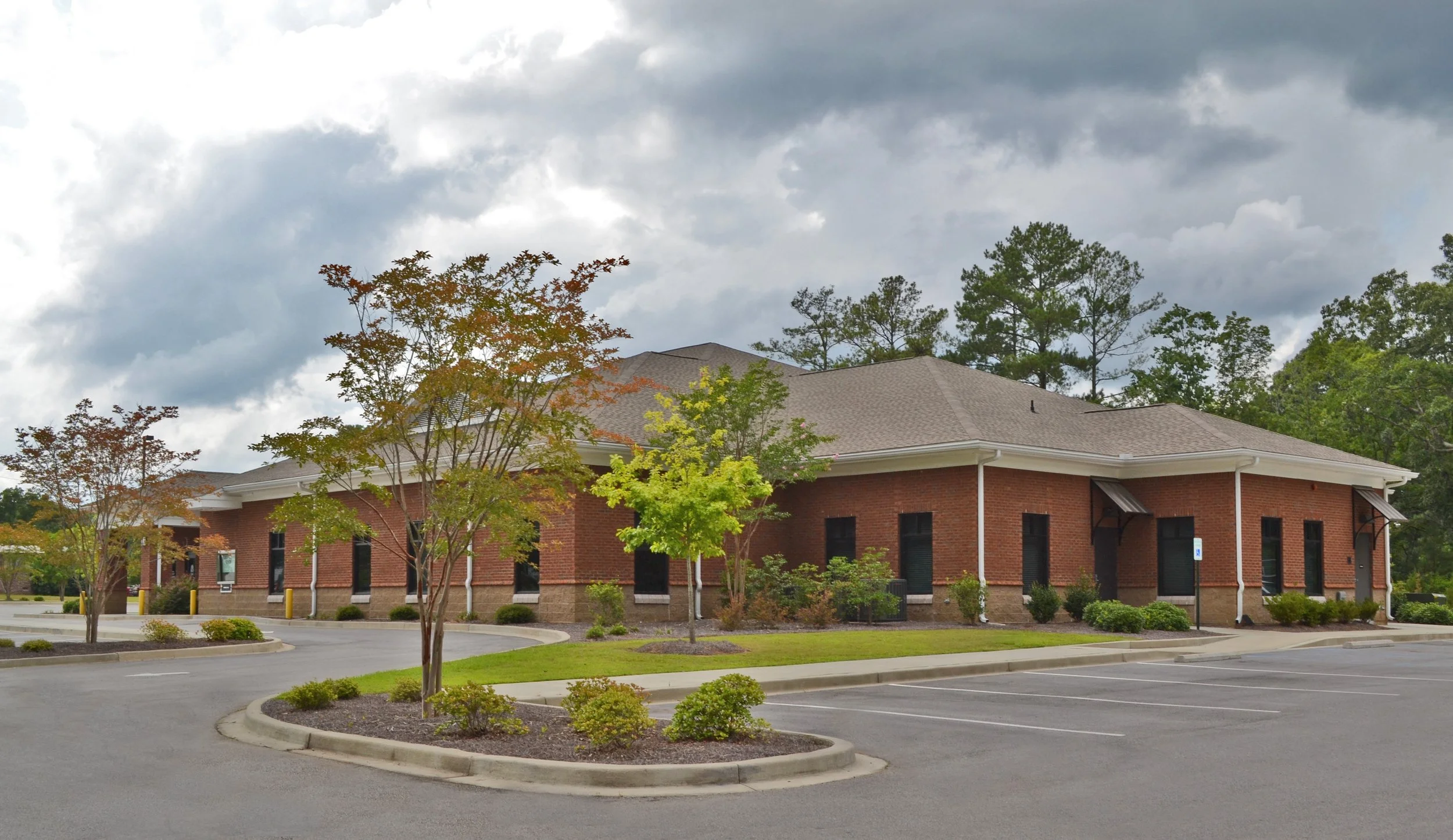 A brick building with a gray shingle roof, surrounded by trees with green and autumn-colored leaves, and a parking lot with a few parking spaces along with small landscape plants. Overcast sky with dark clouds.