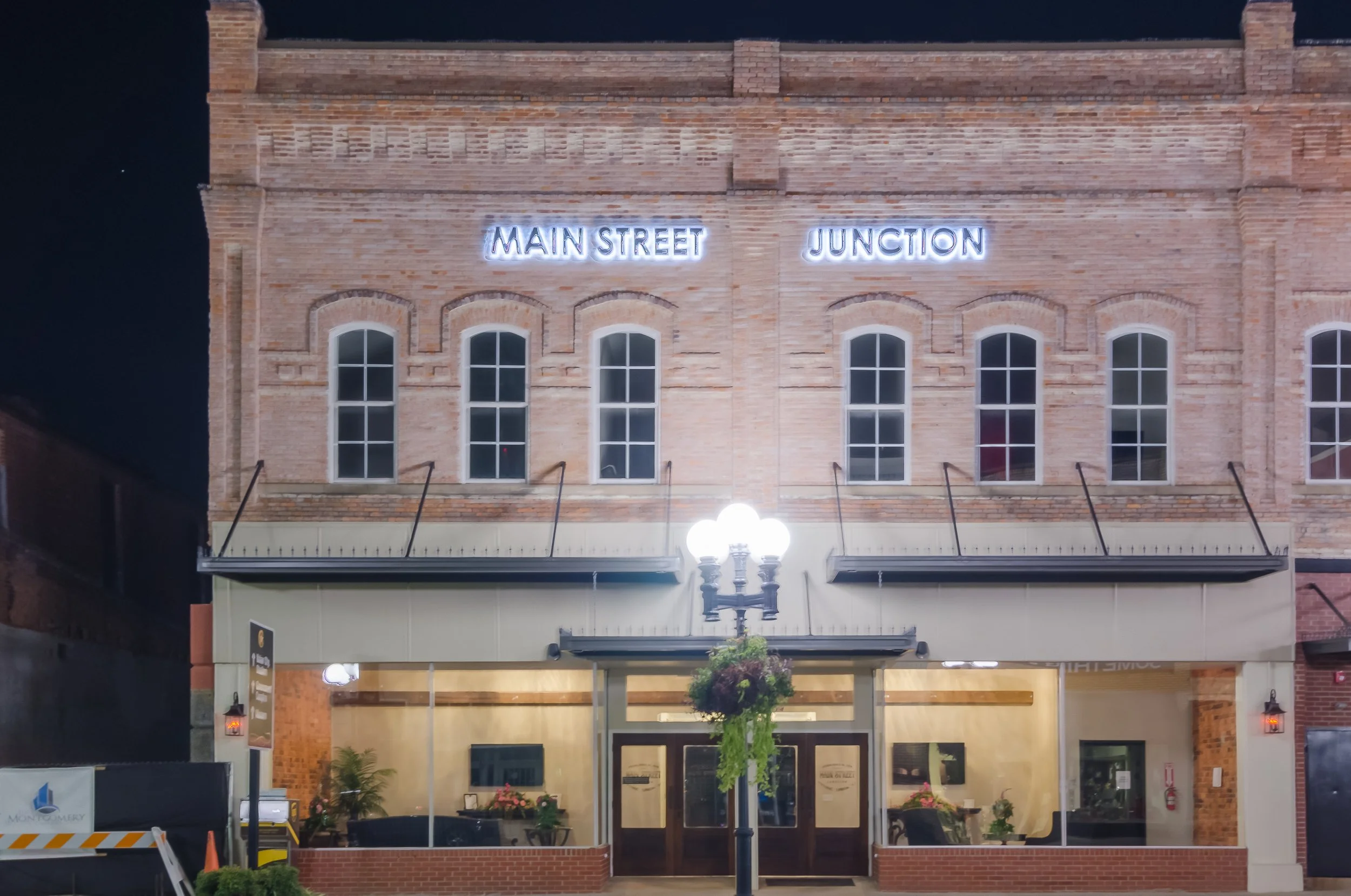 Brick building with illuminated sign reading 'Main Street Junction' above the entrance at night, decorated with hanging plants and outdoor street lamps.
