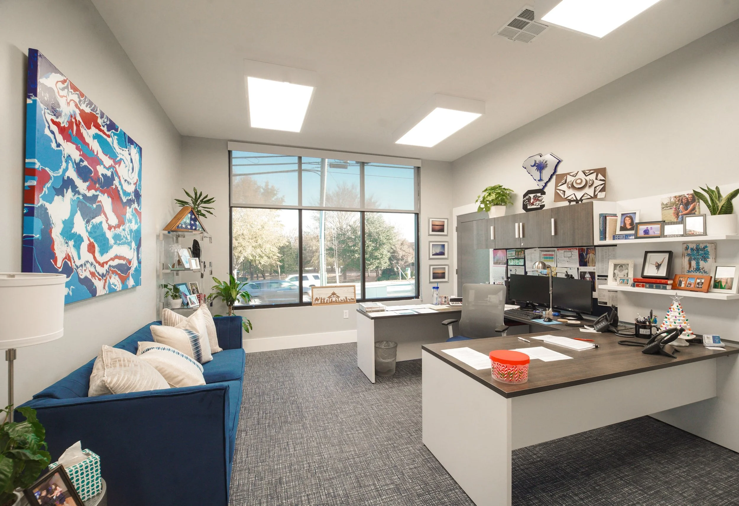 Office interior with a blue sofa, white pillows, large colorful abstract painting on the wall, and a desk with computer monitors and framed photos. Large windows showing trees and a parking lot outside.