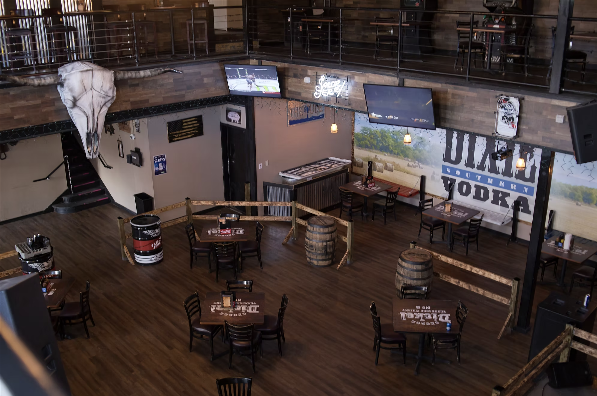 Interior of a Southern-themed restaurant or bar with wooden floors and walls, featuring a mounted animal skull with long horns, multiple television screens showing sports, tables and chairs with betting menus, barrels used as tables, string lights, neon signs, and western decorations.