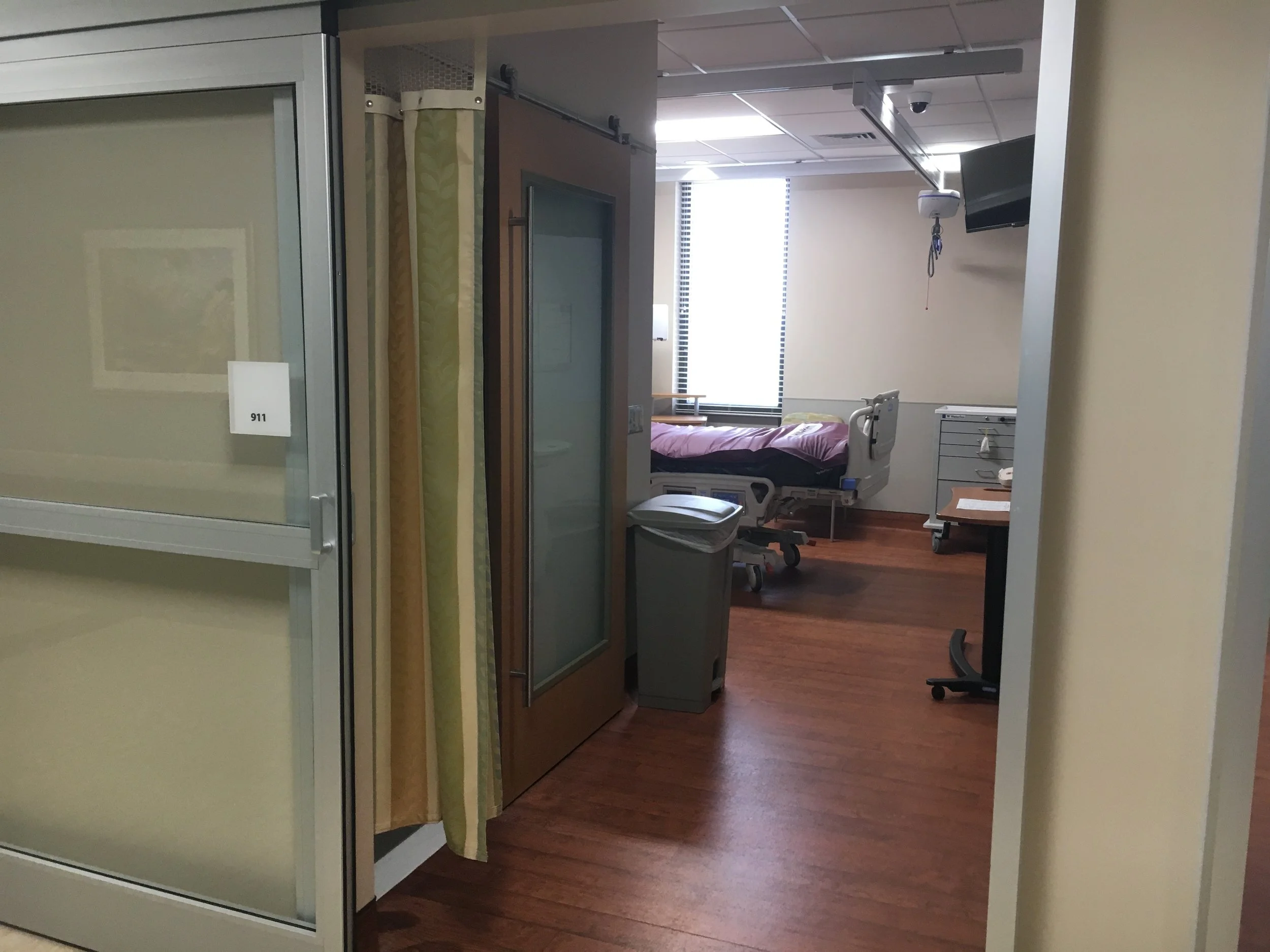 View of a hospital room with an empty bed, medical equipment, and a TV mounted on the wall, seen through a doorway.
