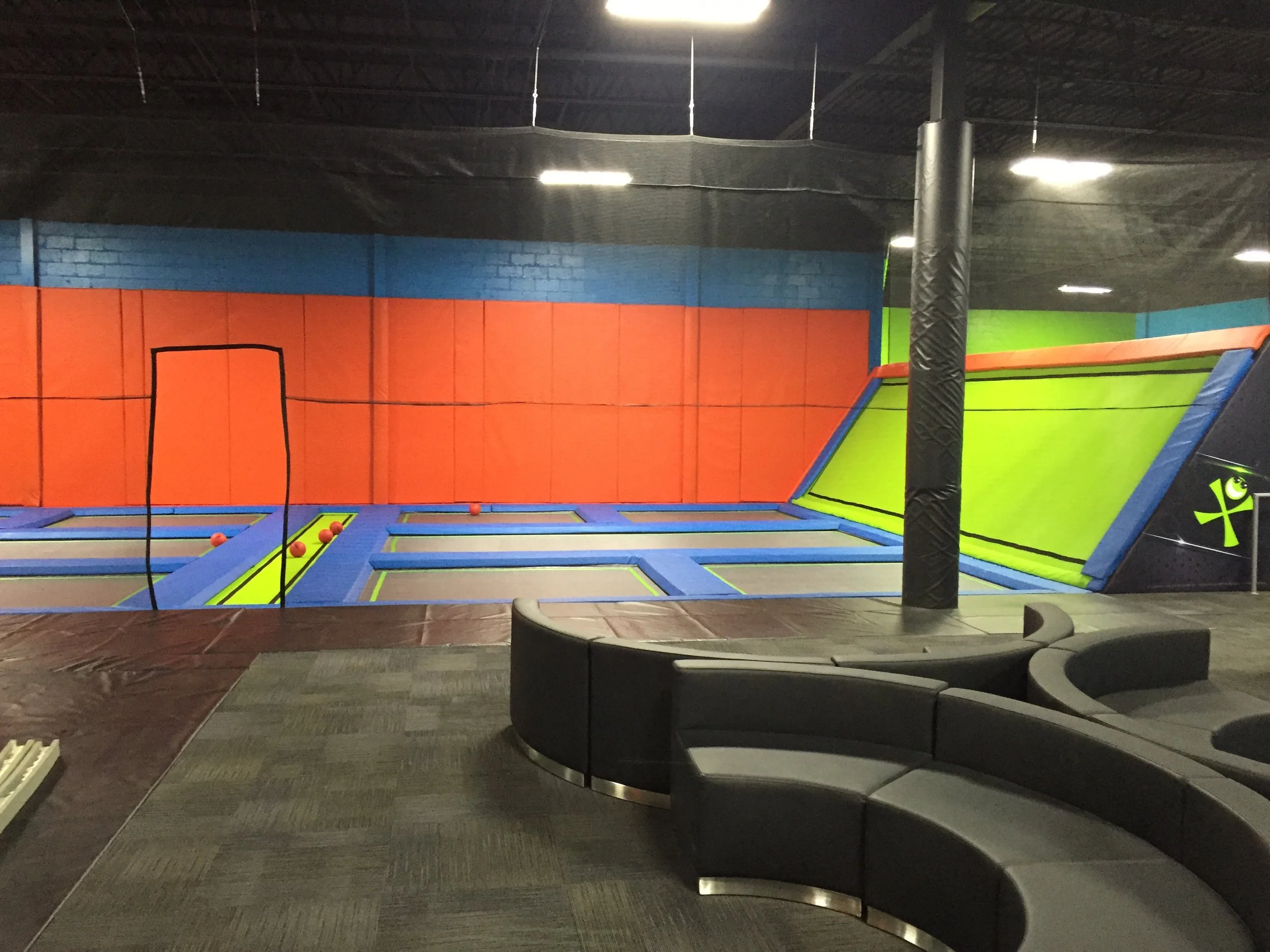 Indoor trampoline park with colorful mats and padded walls. A black pole supports padded cushioning, and a black and green area is visible on the right. The ceiling has black panels with bright lights.