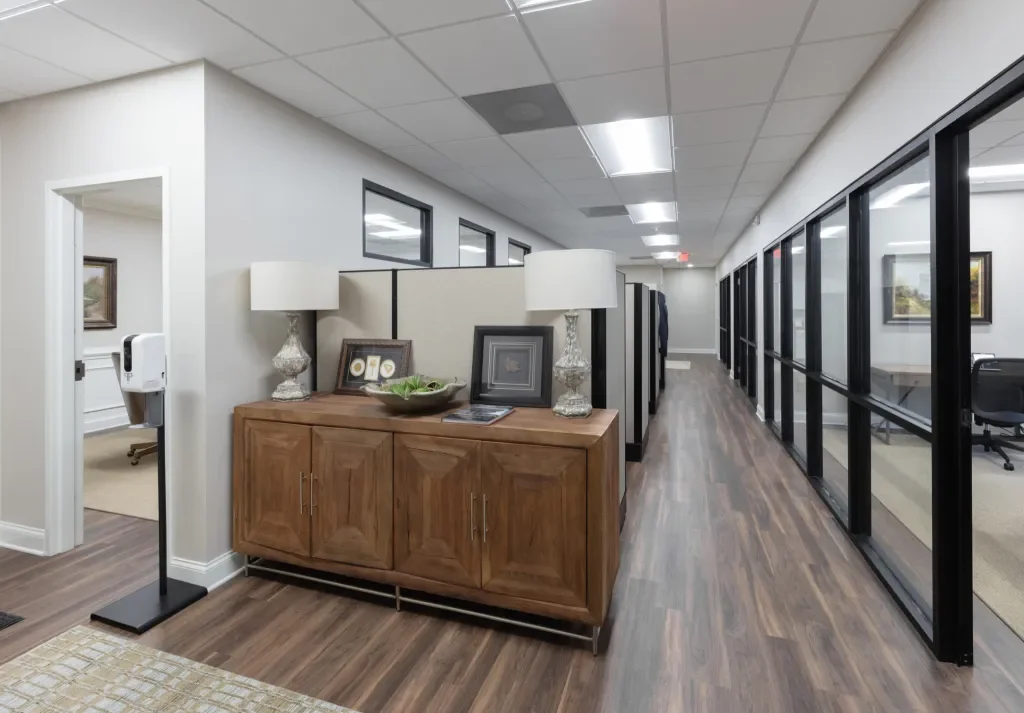 Interior view of an office with a wooden sideboard, two table lamps, picture frames, and decorative items, a hallway with glass walls on the right, and an adjoining room visible through an open doorway on the left.