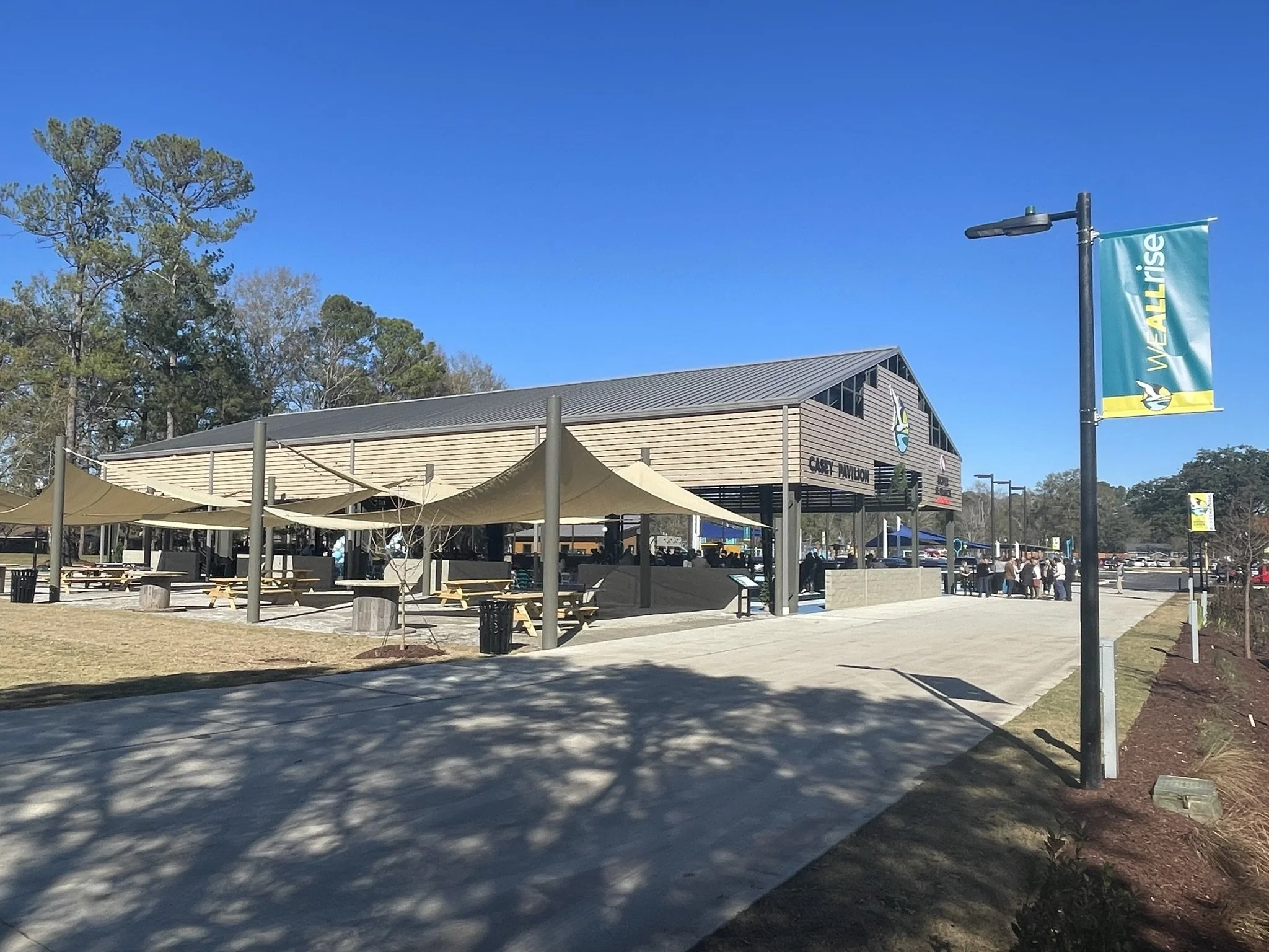 An outdoor scene at a public space with a large modern building labeled 'CASEY PAVILION', a blue sky, tall trees, and a flag that reads 'WEEALL IRISE' with a penguin logo. There are shaded picnic tables and a line of people waiting in the background.