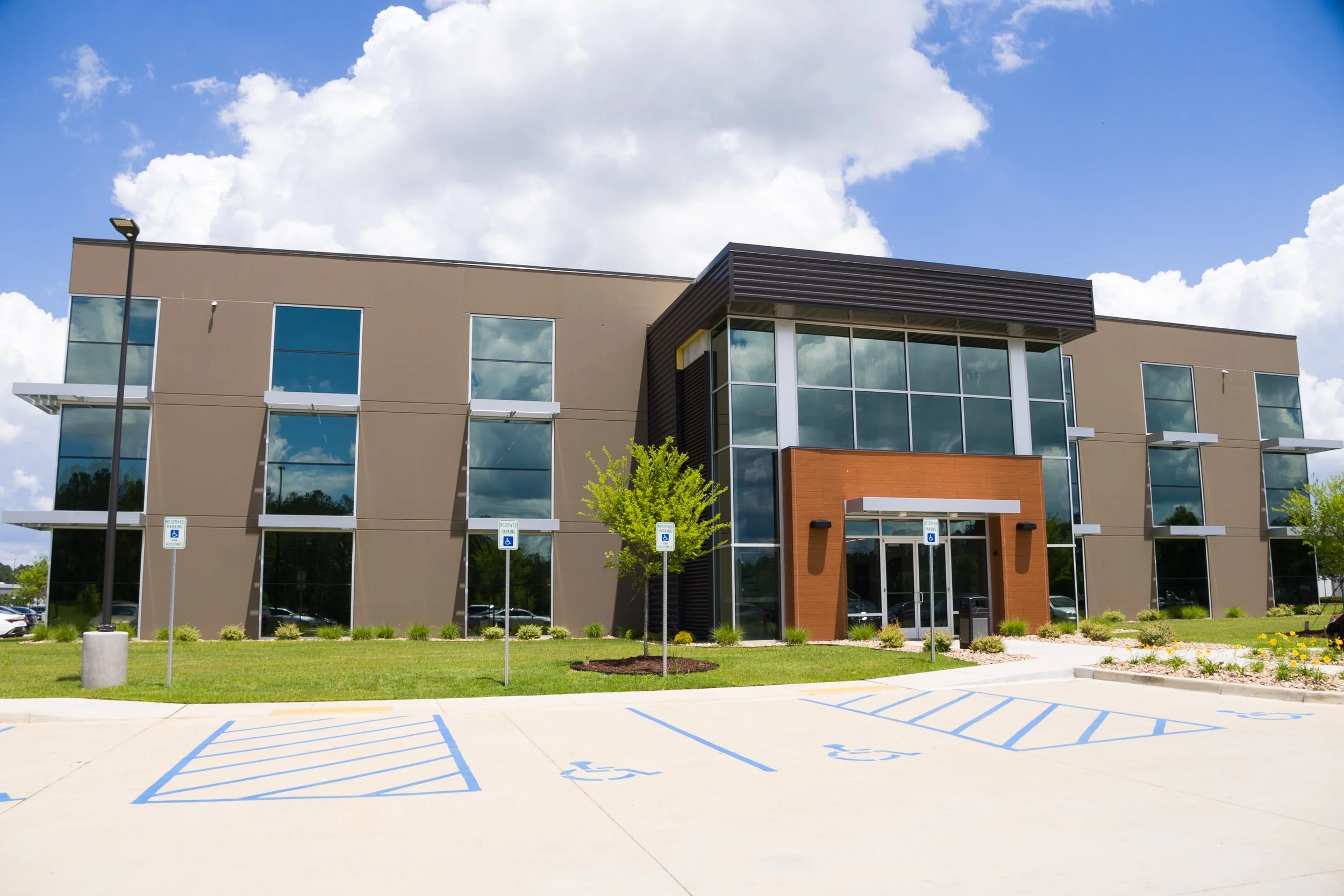 Modern office building with large windows, beige and brown exterior, surrounded by a parking lot with accessible parking spaces, landscaping with small trees and bushes, under a partly cloudy sky.