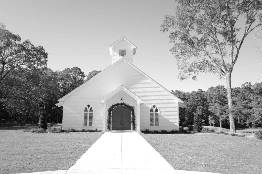 Black and white photo of a small white church with a peaked roof, a small bell tower, arched windows, a wooden door with a wreath, and a sidewalk leading to the entrance, surrounded by trees.