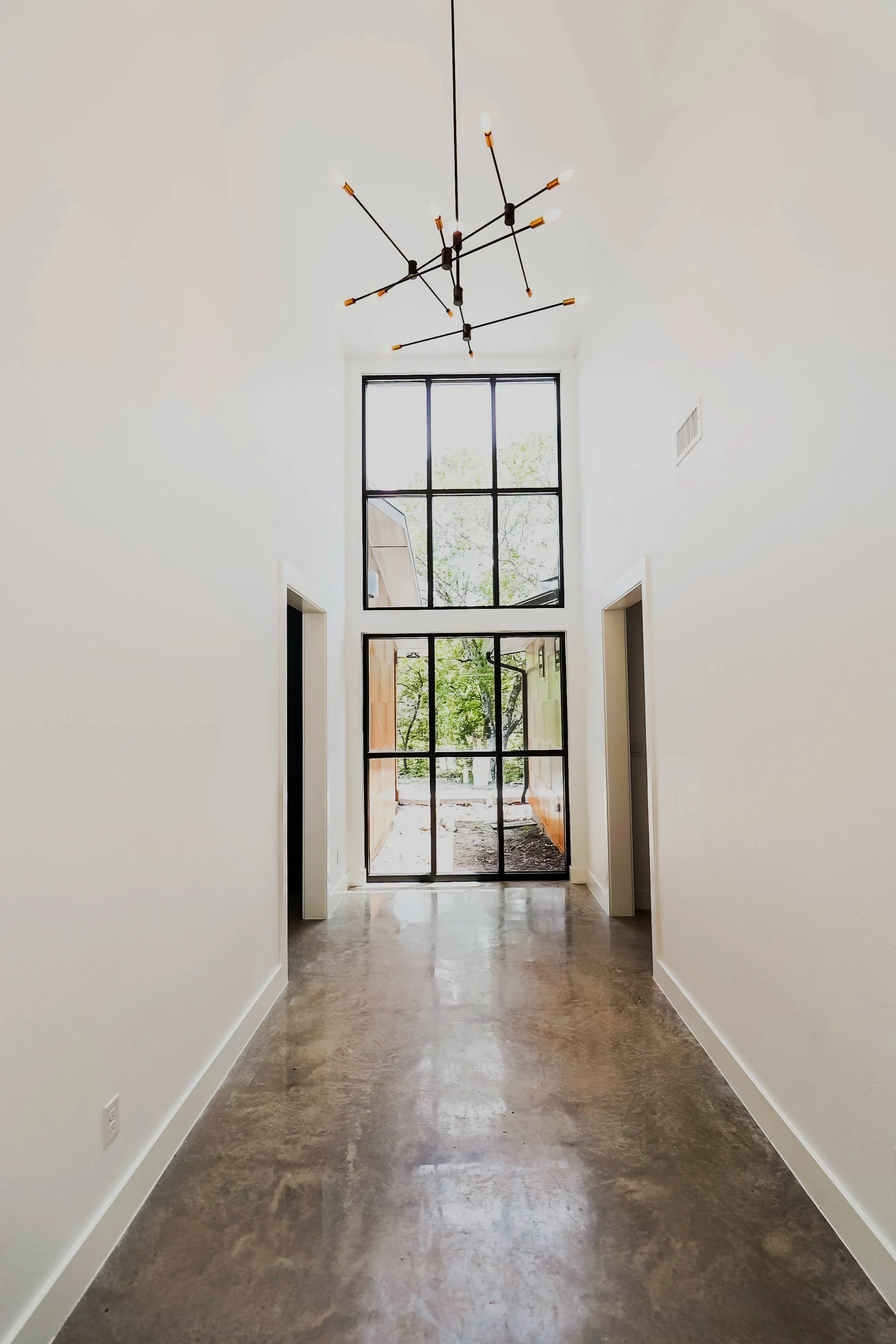 Bright hallway with large window at end, white walls, polished concrete floor, and modern ceiling chandelier.