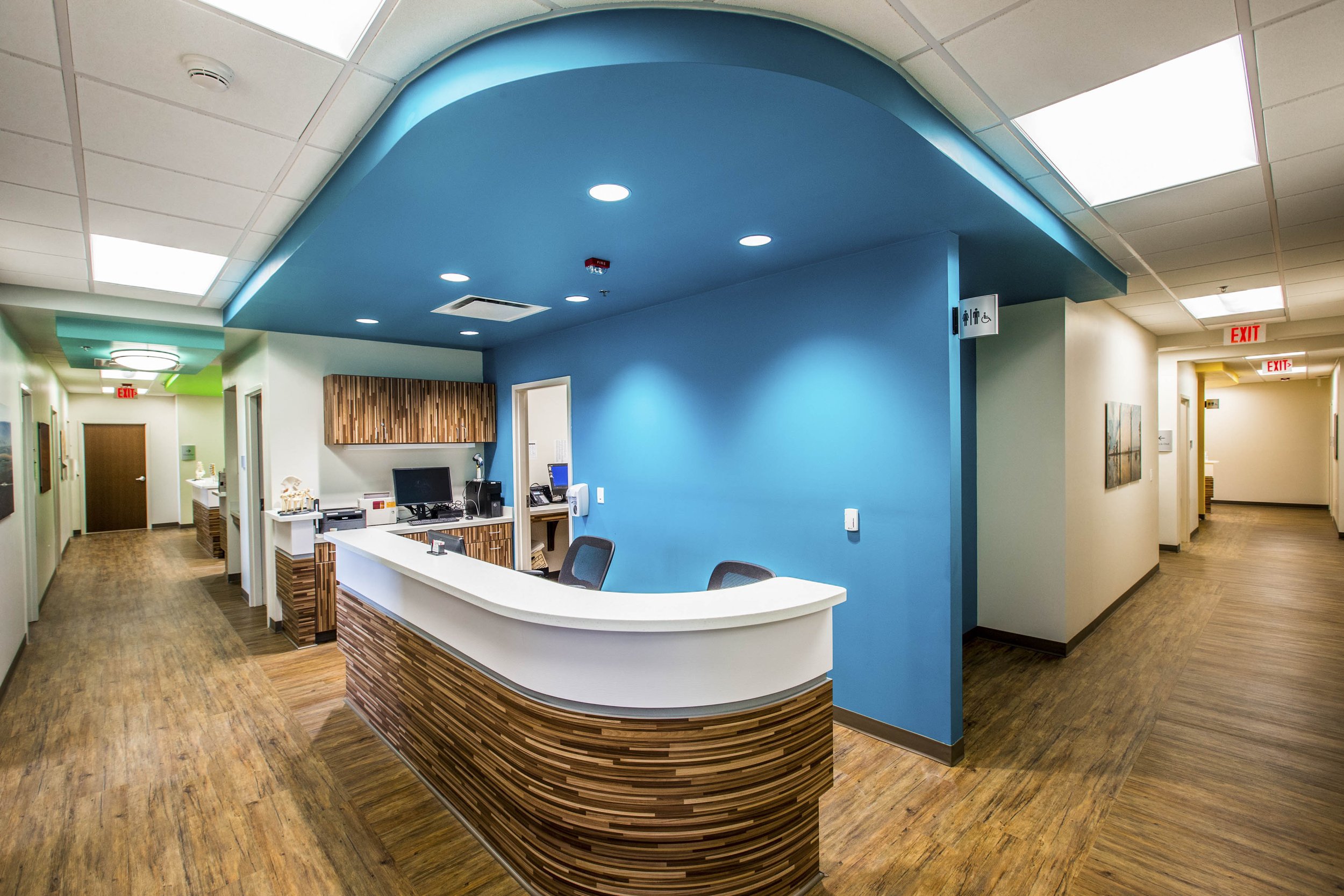 Hospital reception area with a curved wooden front desk, a blue accent wall, and various signs indicating restrooms and exits. Ceiling with fluorescent lighting and wood flooring.