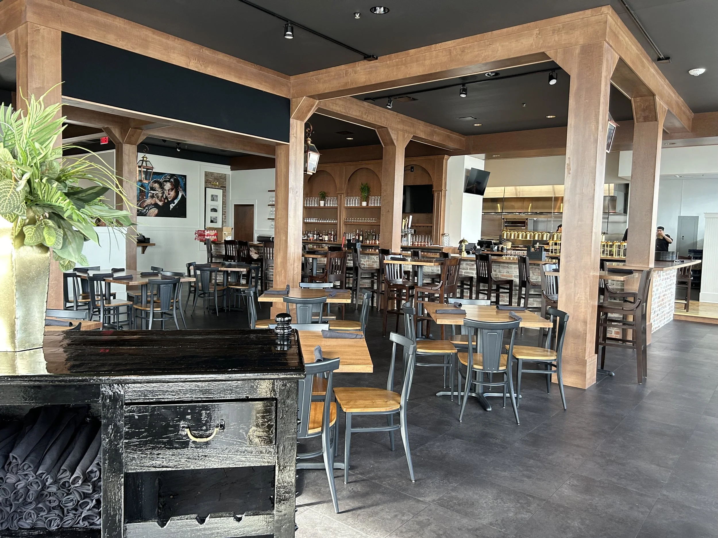 Interior of a restaurant or bar with wooden beams and columns, tables and chairs arranged for dining, a bar area with glassware, and artwork on the wall.