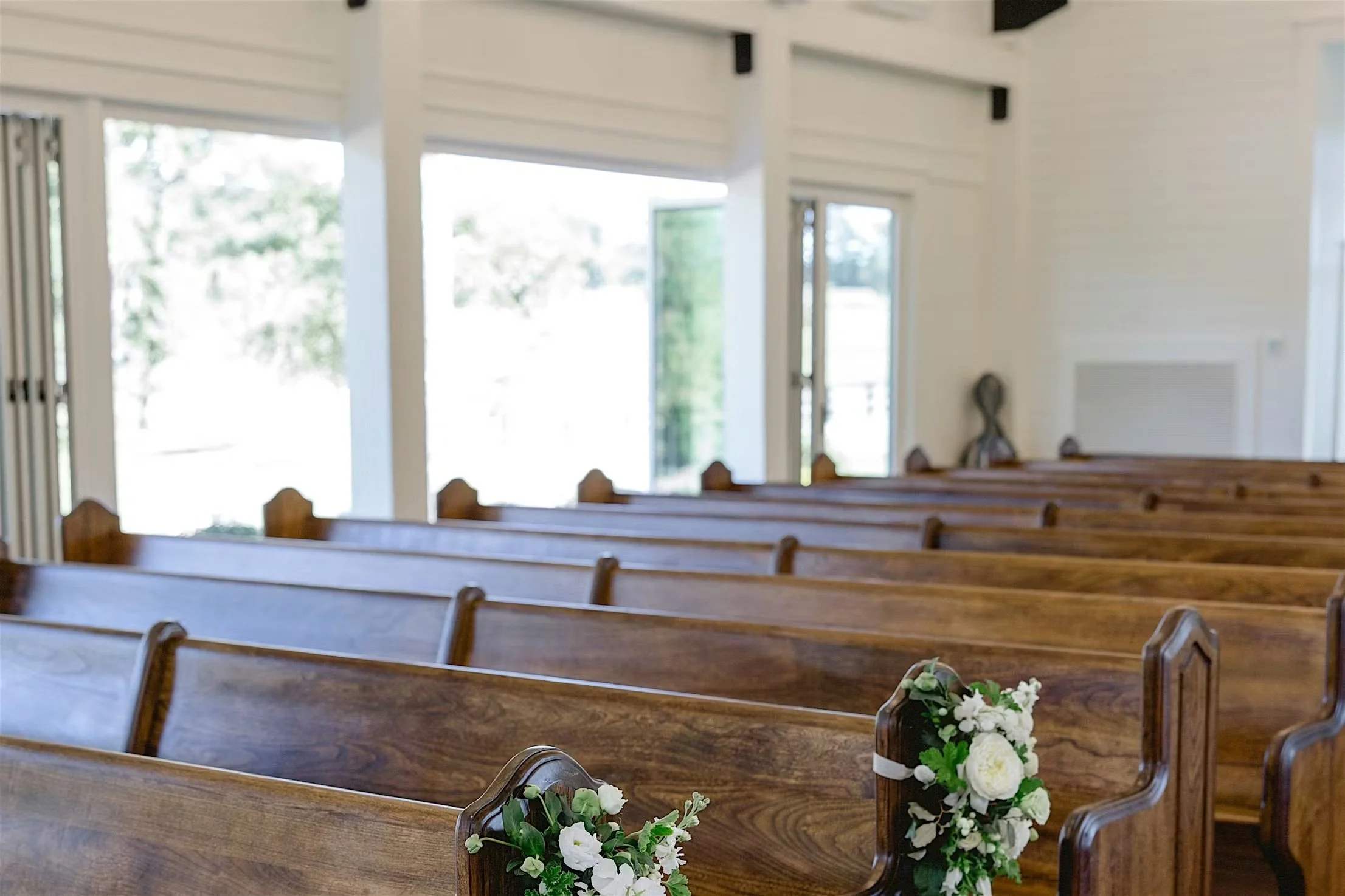 Interior of a church or chapel with wooden pews decorated with white flowers, large windows letting in natural light, and a simple, bright decor.