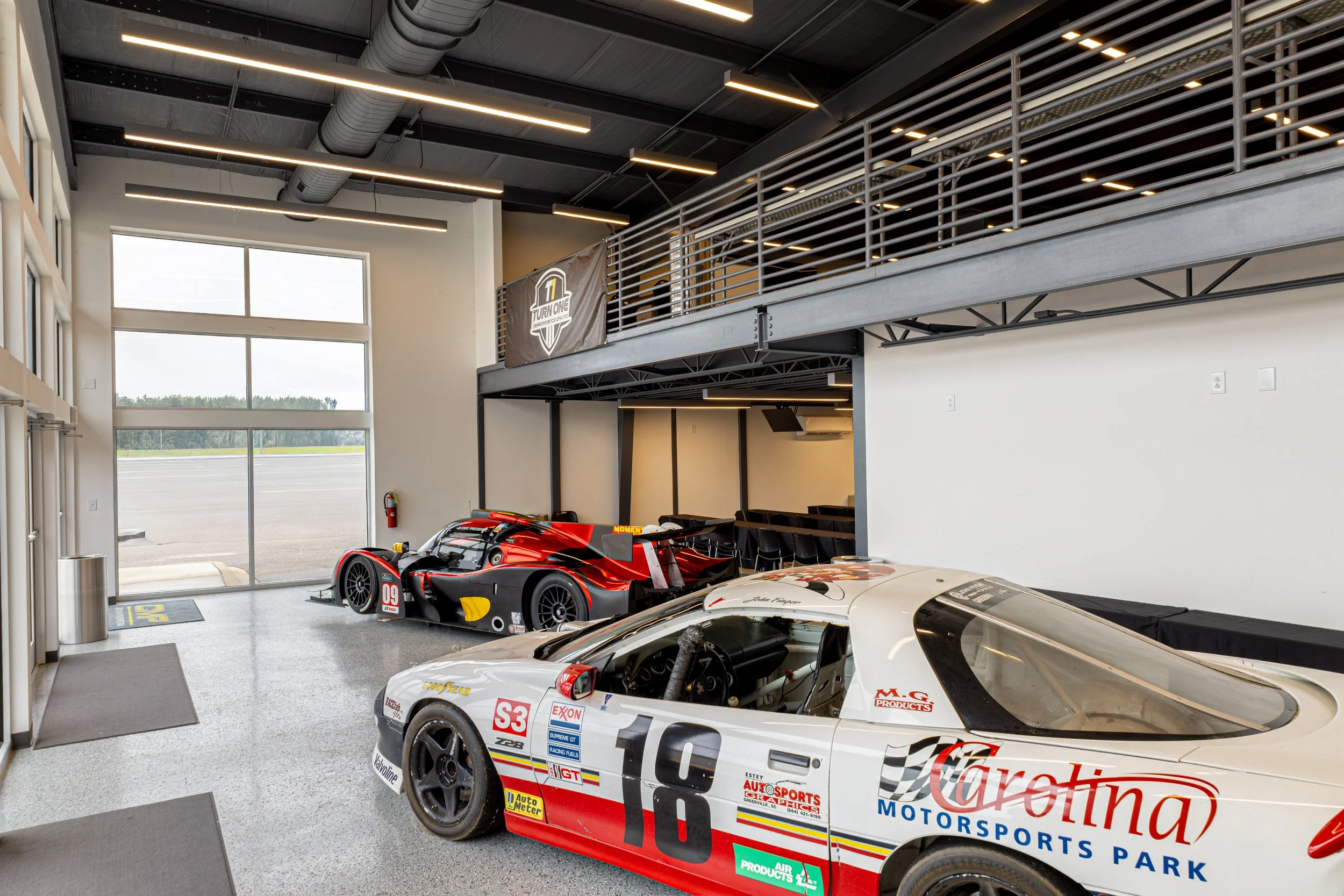 Interior of a race car garage with two race cars parked inside; one white with sponsor logos and the other red and black. Large windows at the back of the garage reveal an airstrip outside. Industrial ceiling lights and ventilation ducts are visible.